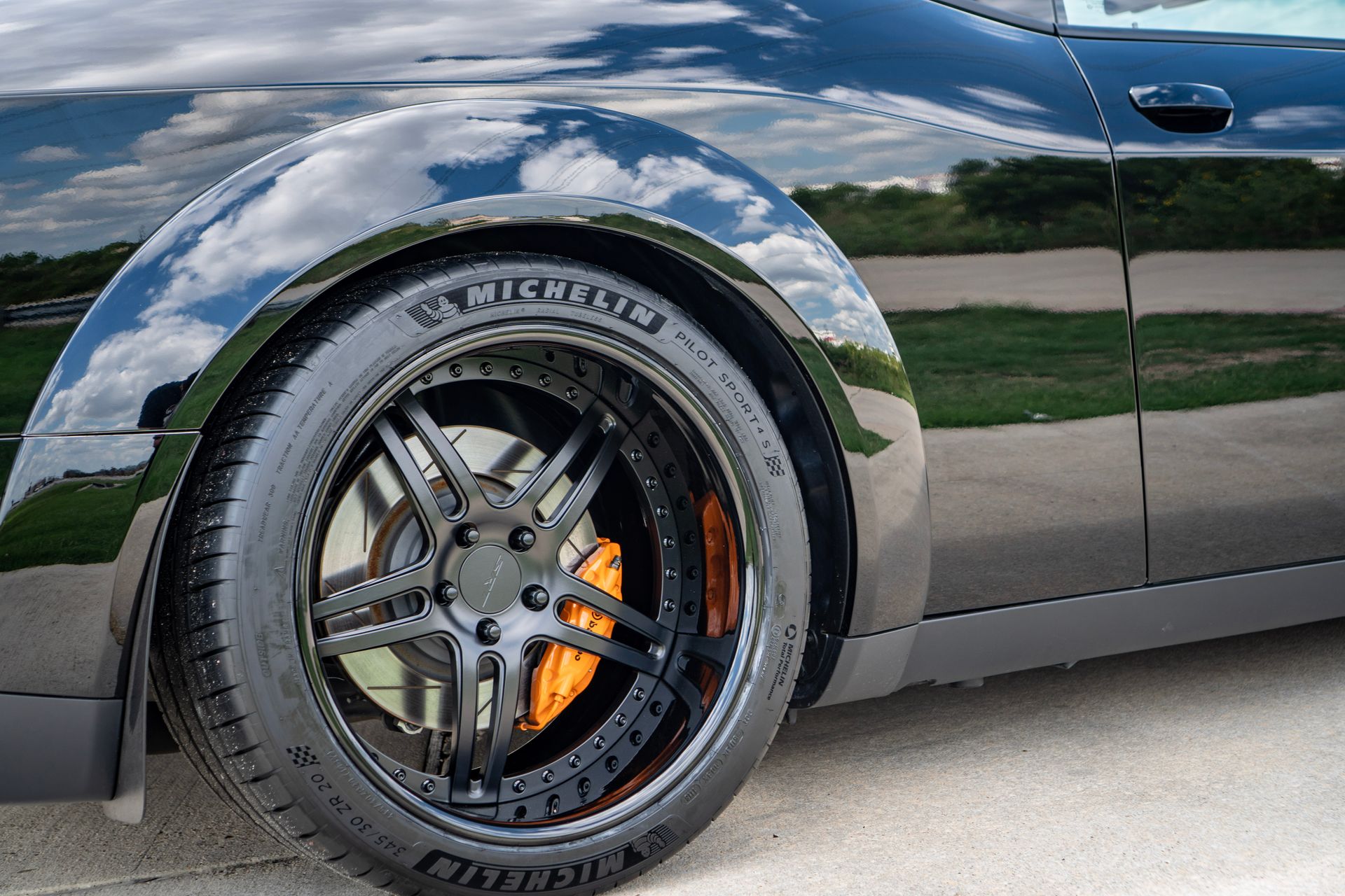 Black car's front tire and wheel, orange brake caliper, with a shiny reflection of the sky.