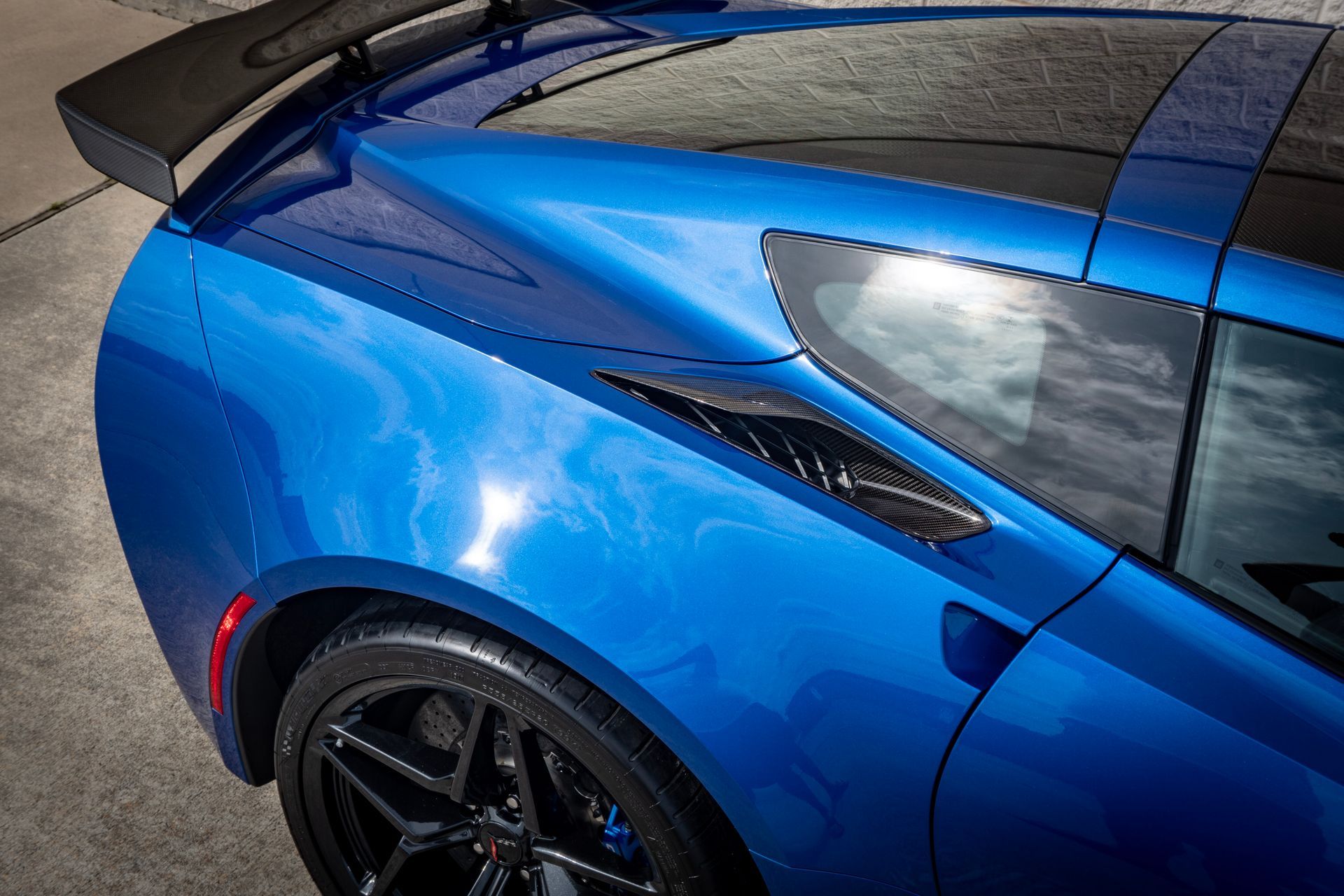 Blue sports car's rear, with black spoiler, vent, and wheel, parked on concrete.