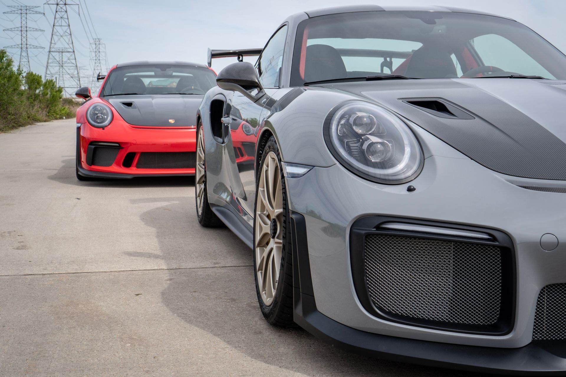 Two sports cars, gray and red, parked on a paved road. Gray car is in front.