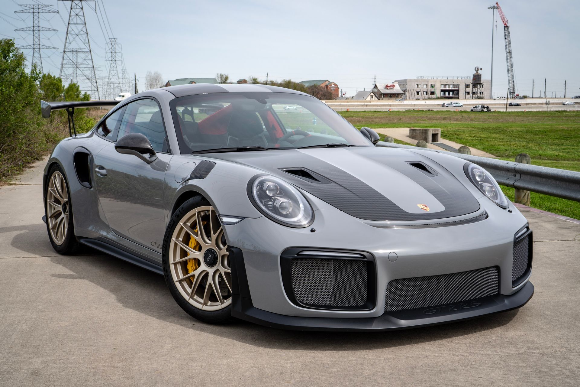 Gray Porsche 911 GT2 RS sports car parked on asphalt, gold wheels, black racing stripe, outdoors.