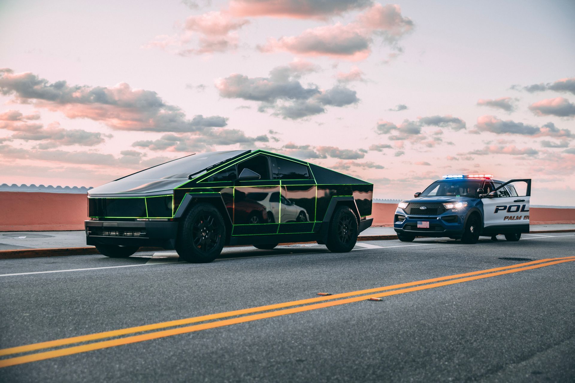A black Tesla Cybertruck with neon green trim is pulled over by a police SUV on a bridge at sunset.