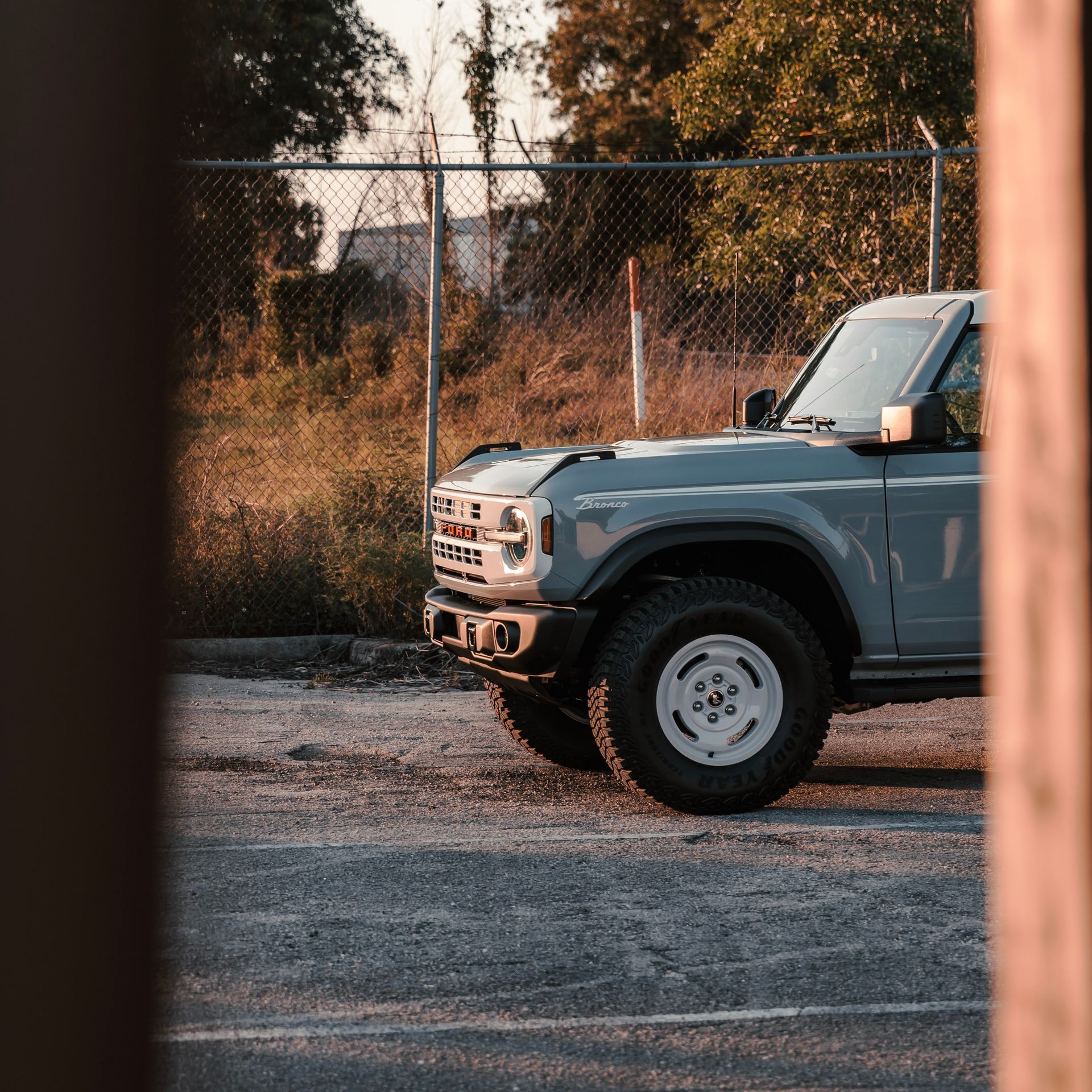 Gray Ford Bronco parked in a gravel lot with a fence and trees in the background.