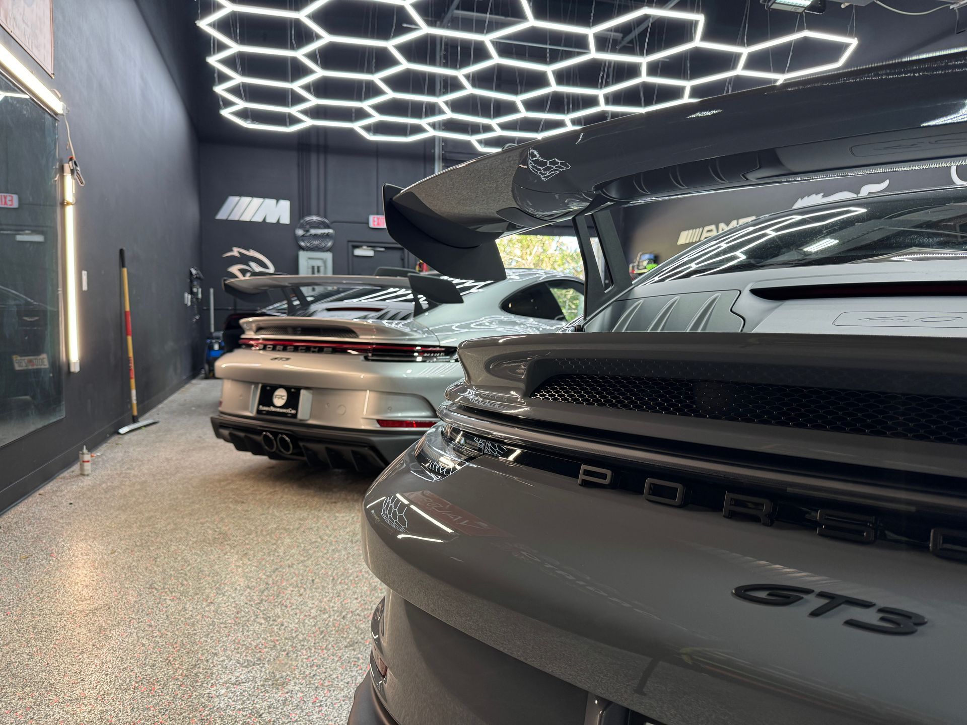Two grey Porsche GT3 sports cars in a garage under hexagonal lights.