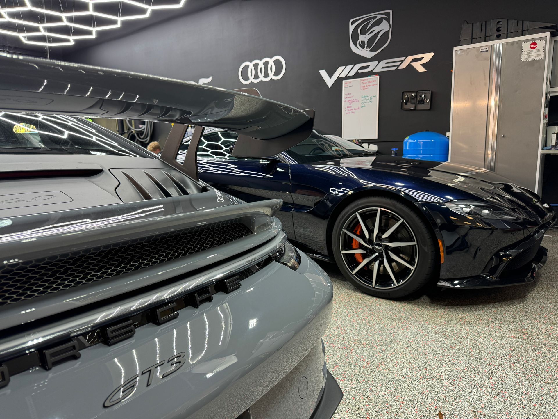 Two sports cars, one gray and one blue, parked in a garage with car logos on the wall.