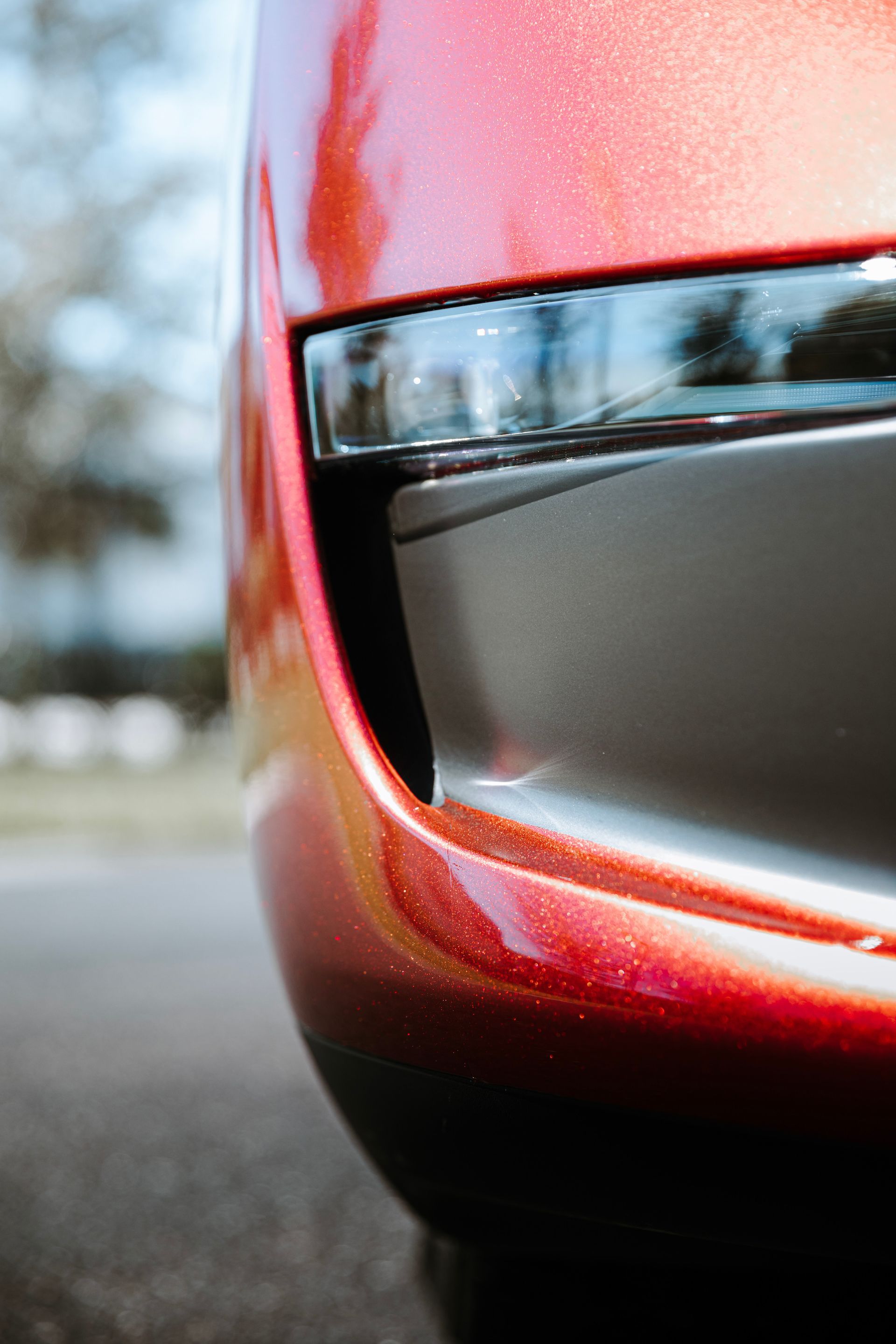 Close-up of a red car bumper with a black trim detail, on asphalt, with blurred background of trees.