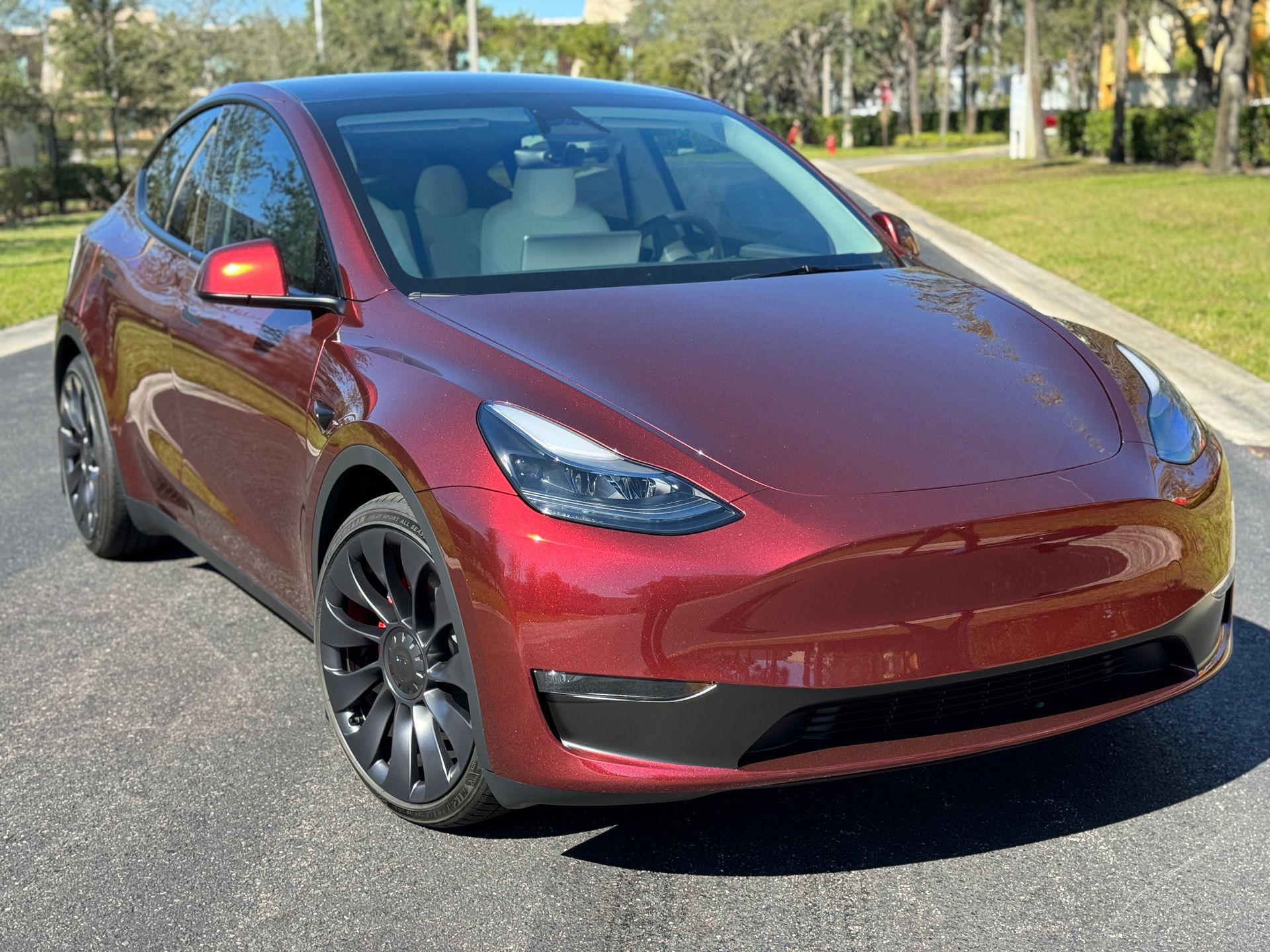 Red Tesla Model Y car parked on a road. Black rims, black roof, sunny day.