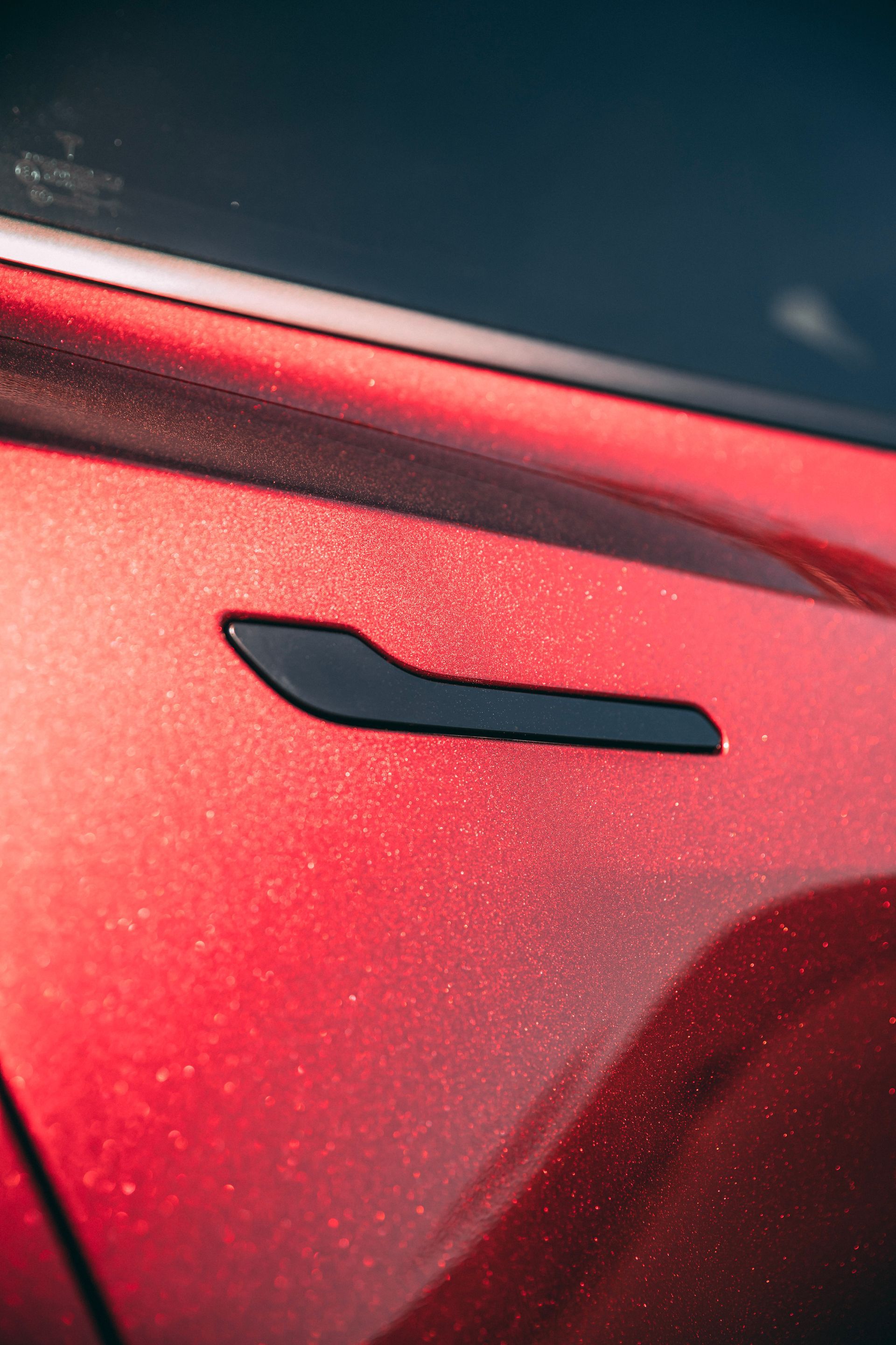 Close-up of a red car door with a black handle, reflecting sunlight.