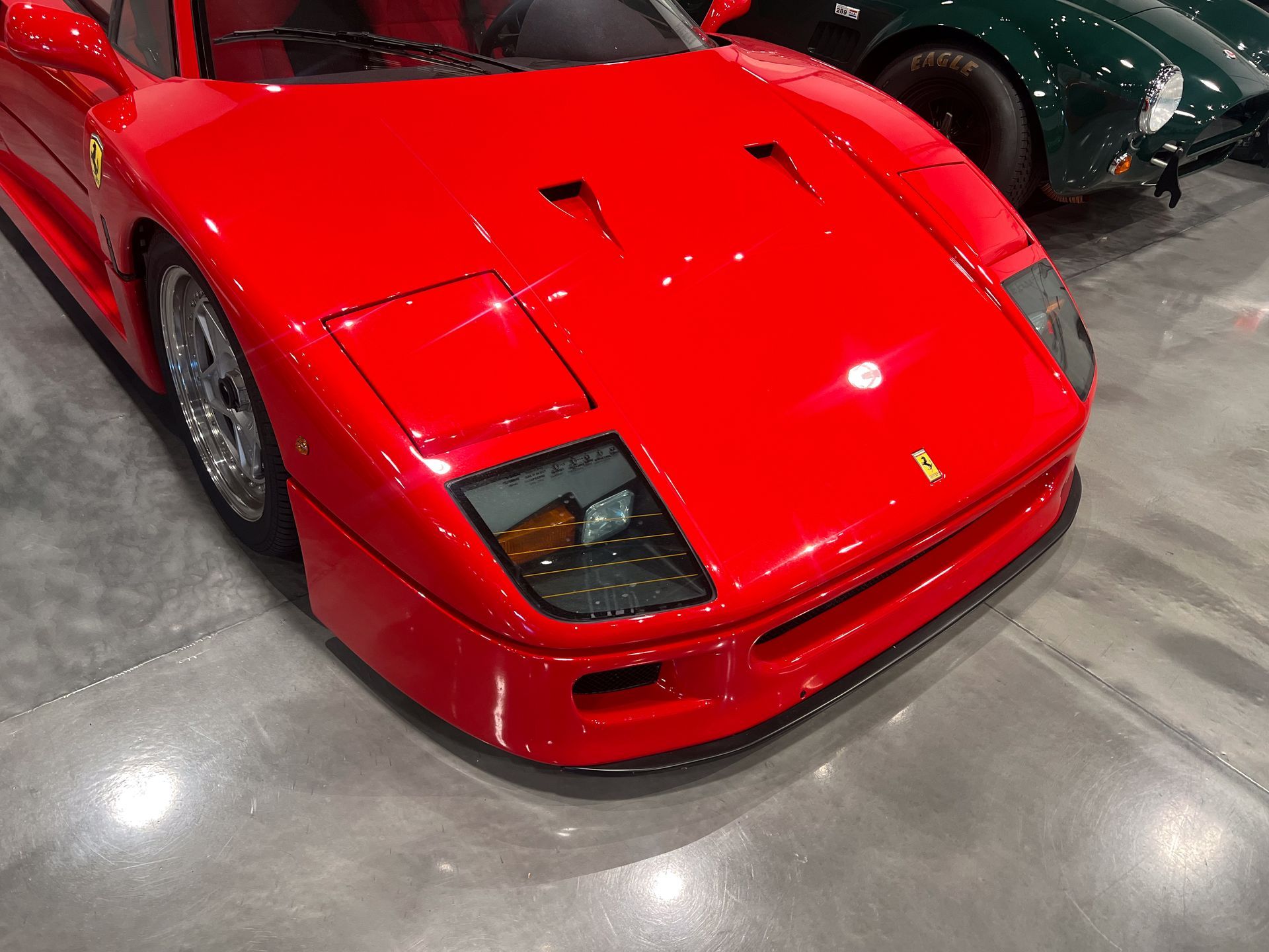 Red Ferrari F40 sports car, front view, in a showroom, with a green car visible in the background.