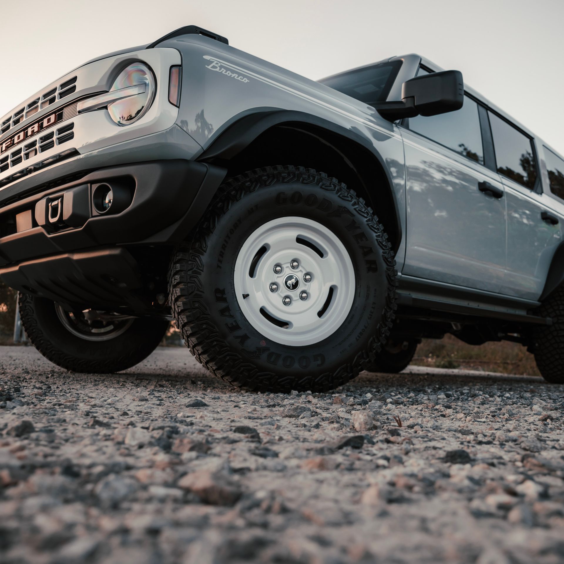 Gray Ford Bronco off-road vehicle with white wheels on a gravel road, low angle view.