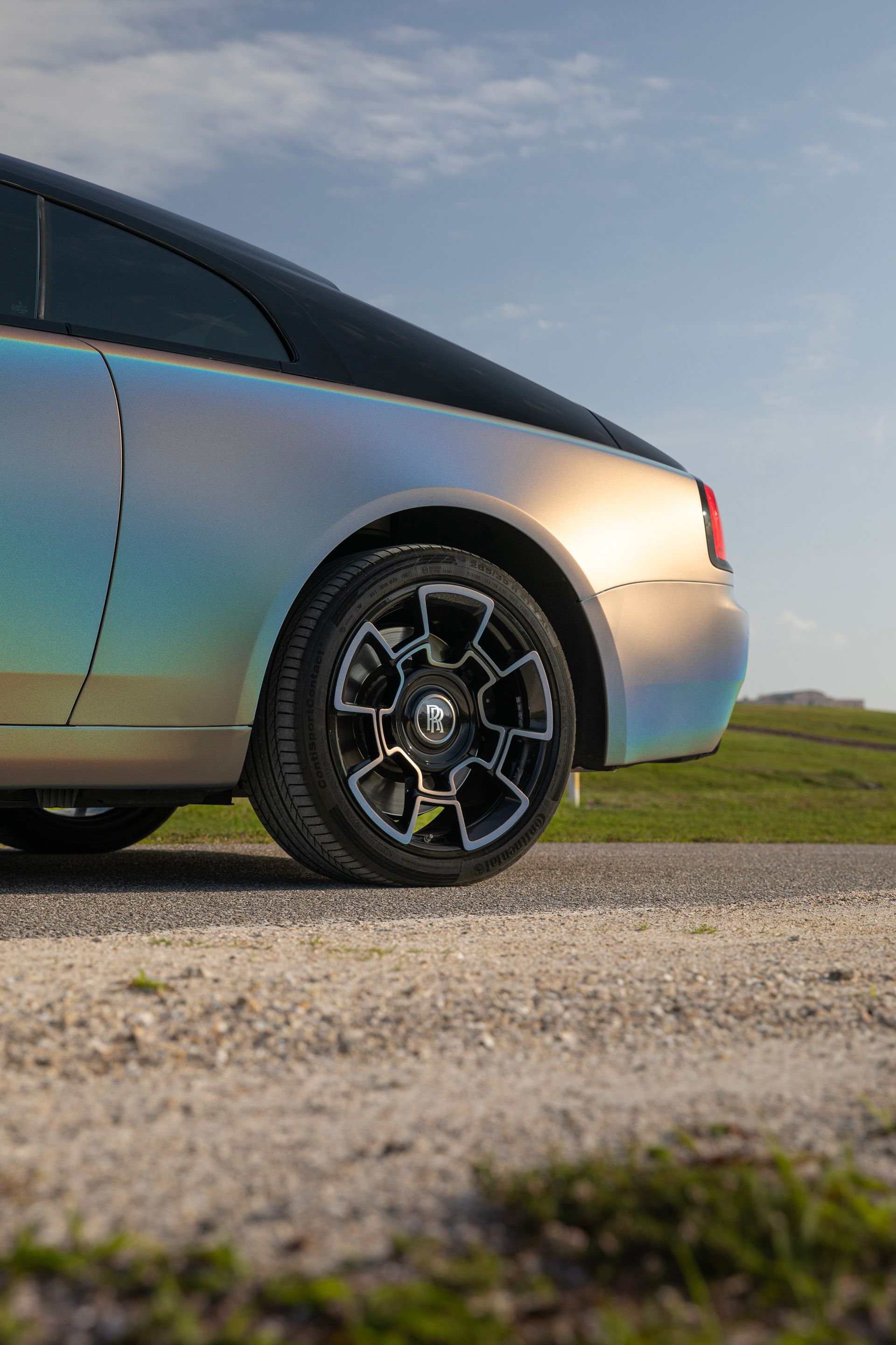 Rear view of iridescent Rolls-Royce coupe parked on gravel road with black and silver rims.
