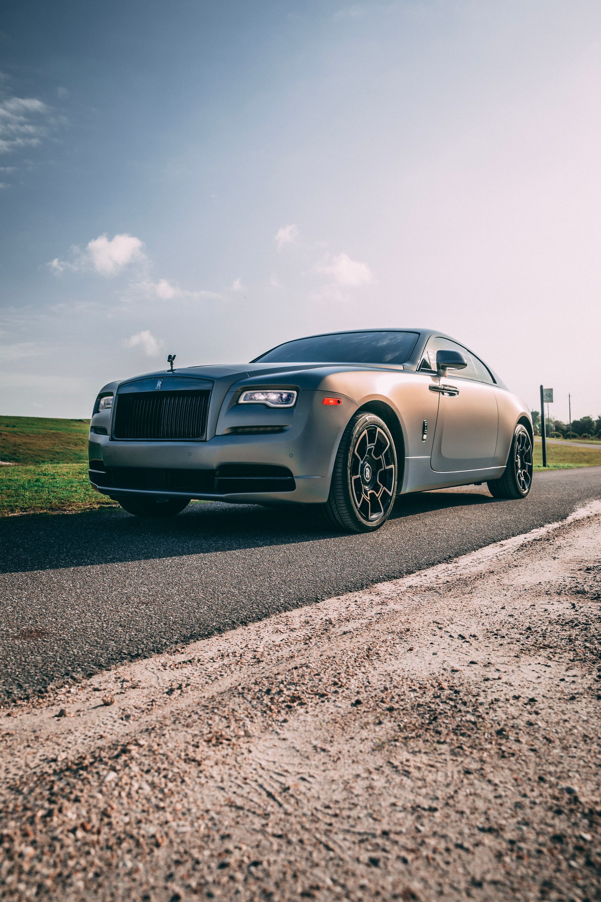 A grey Rolls-Royce coupe parked on a gravel road, with a blue sky background.