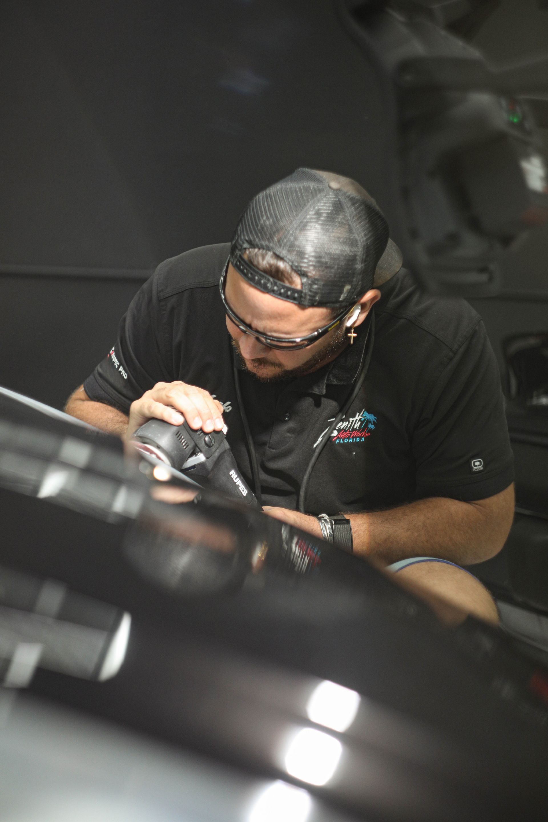 Person polishing a black car with a power tool, wearing safety glasses and a black cap, in a workshop.