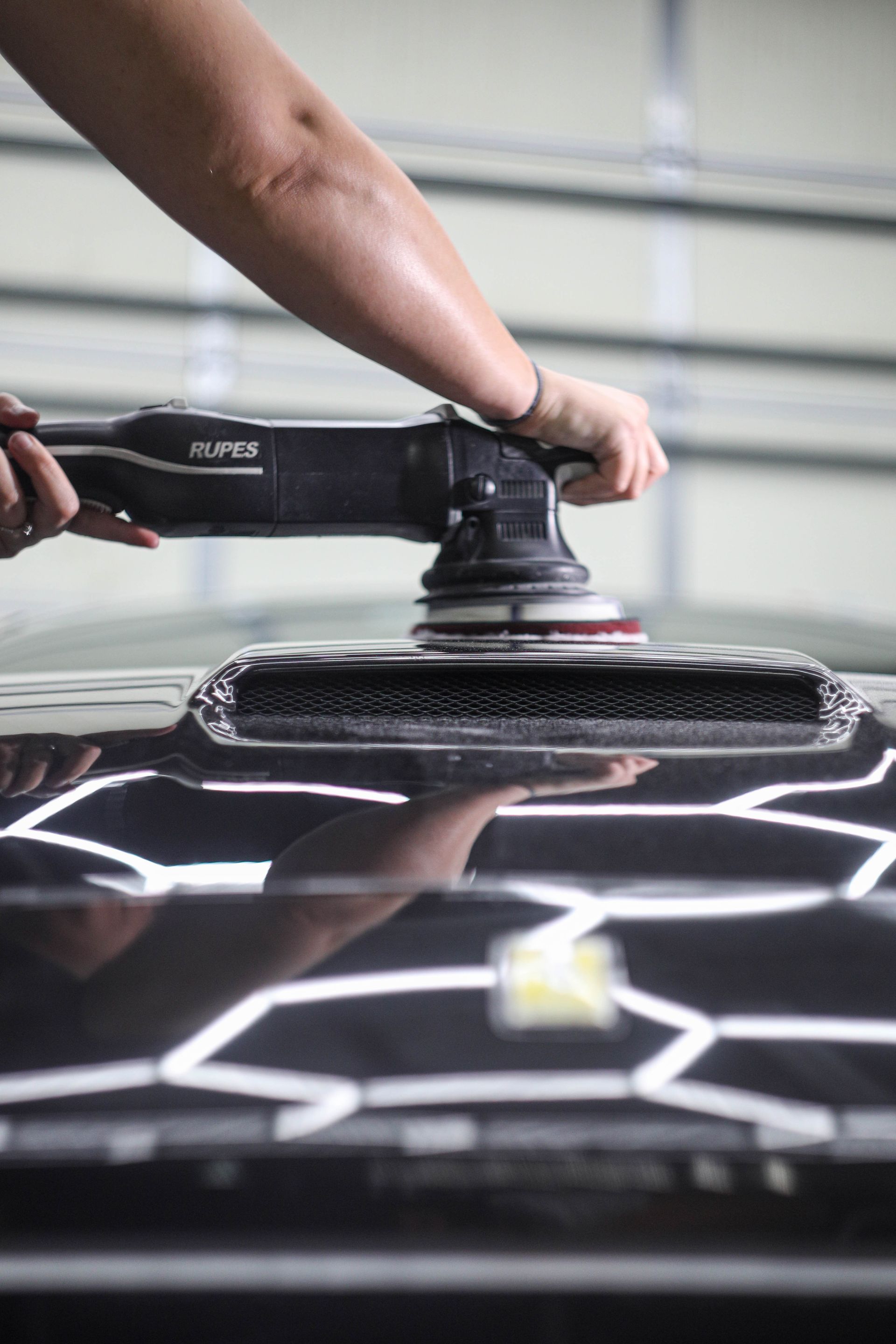 Person using a polisher on a black car hood in a garage, reflecting bright lights.