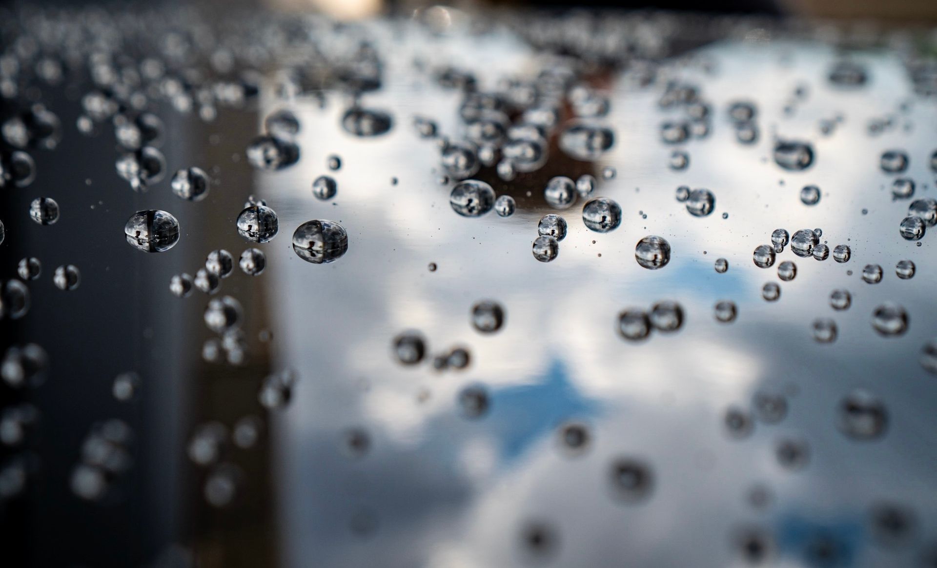 Water droplets on a reflective surface, sky and clouds reflected within them.