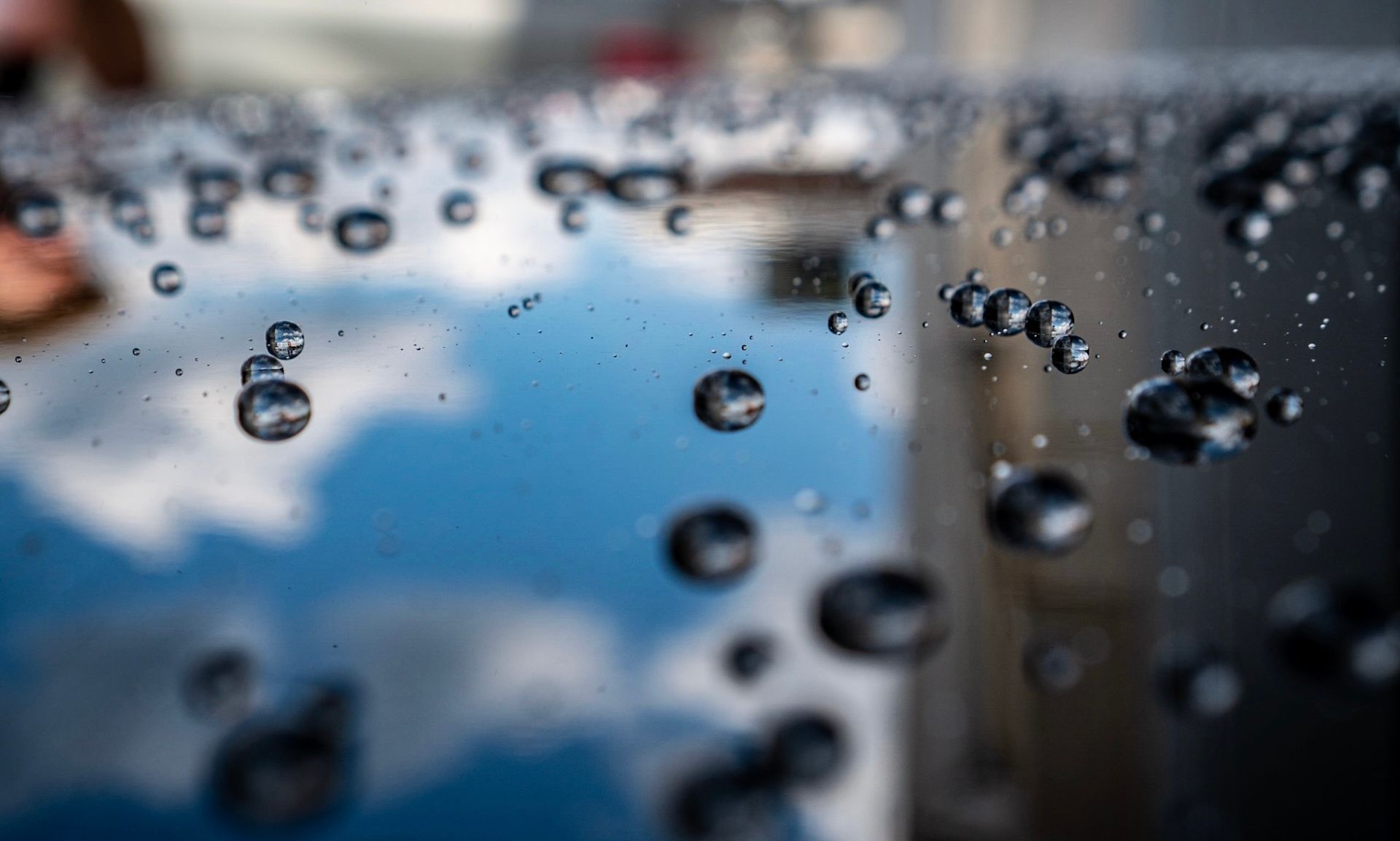 Water droplets on a shiny, black surface, reflecting a sky with clouds.