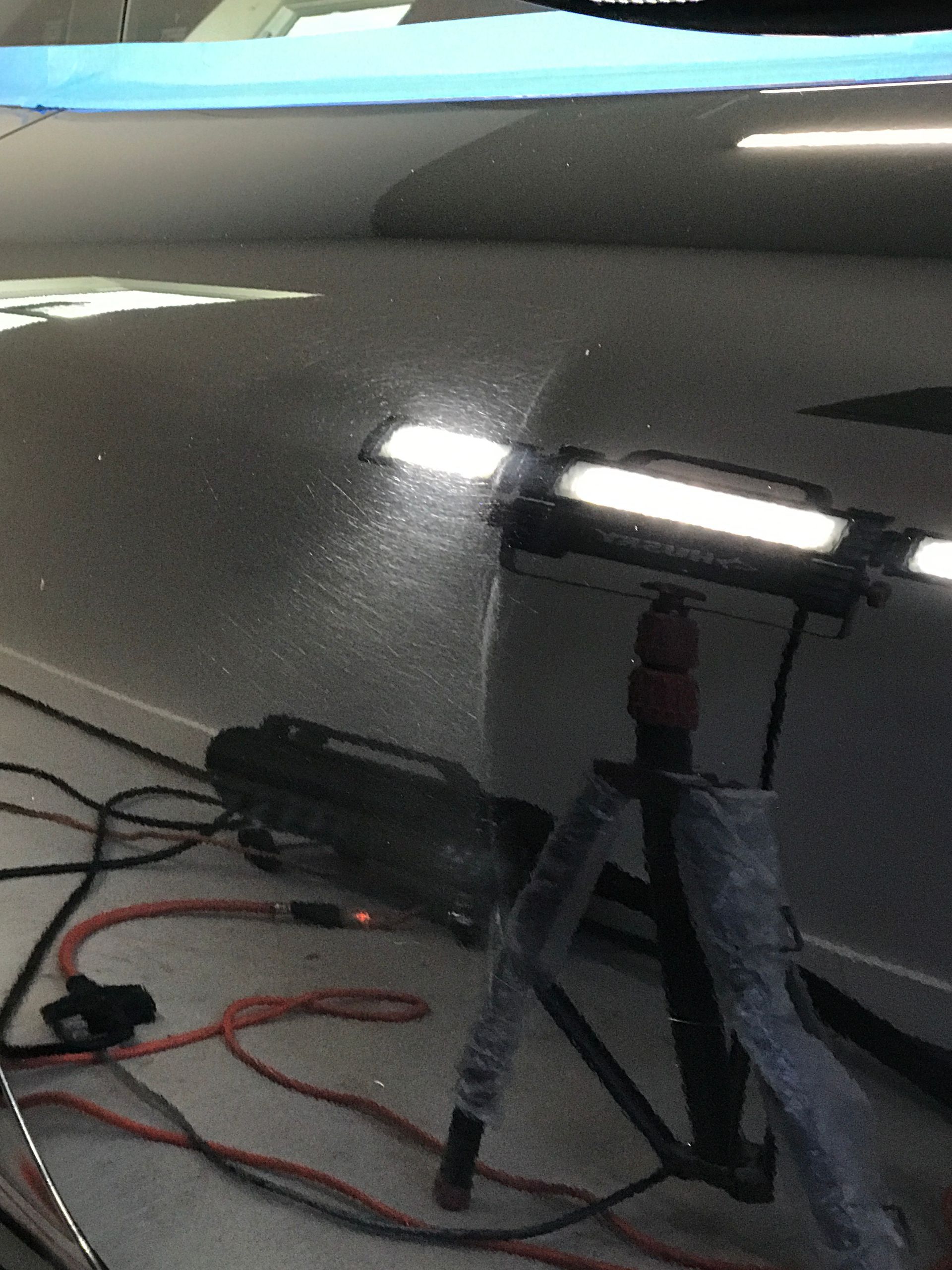 A light shines on a car's surface, reflecting imperfections; cords and a tripod are in the foreground.