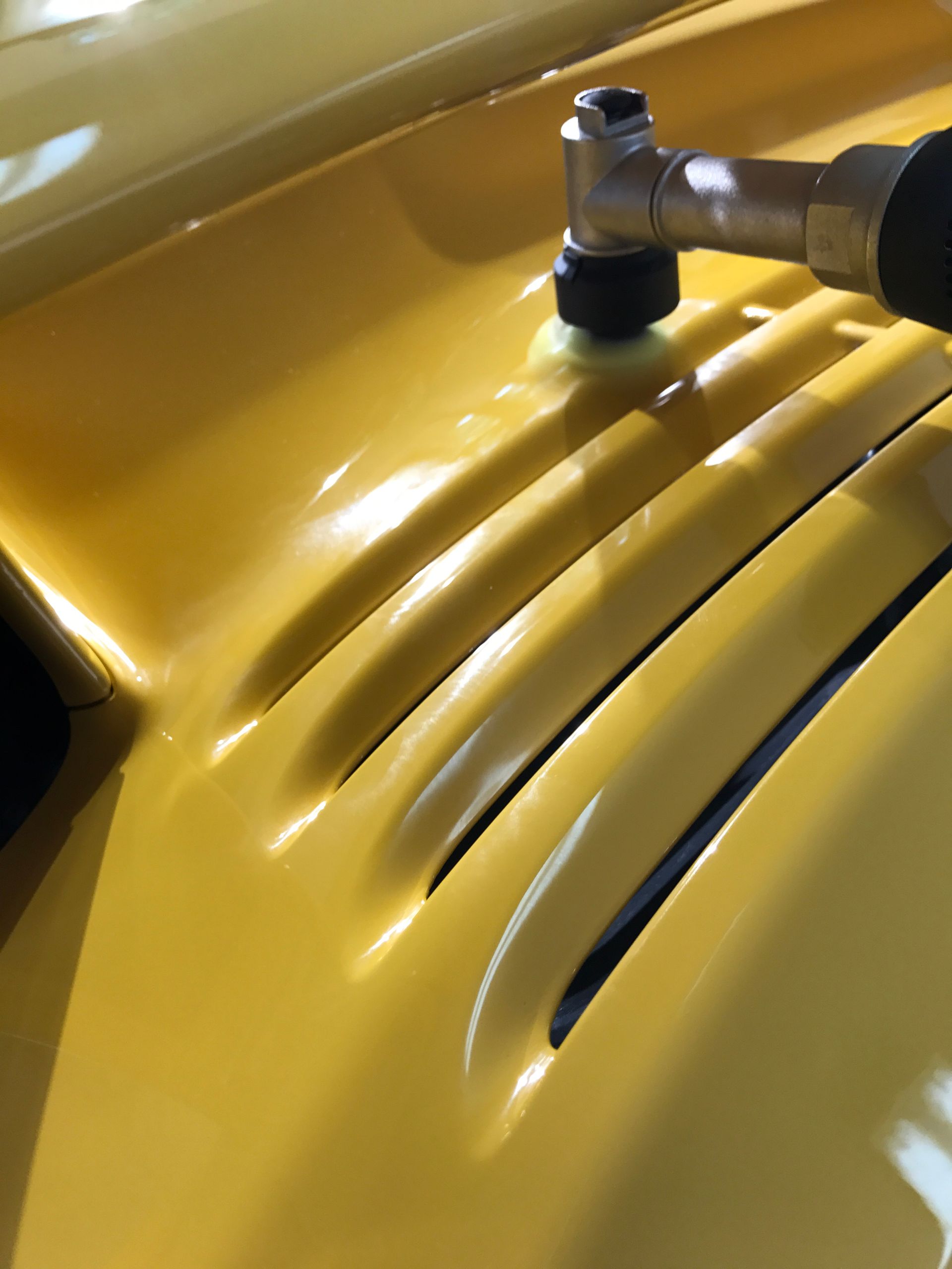 Yellow car hood being polished with a small, motorized buffer.