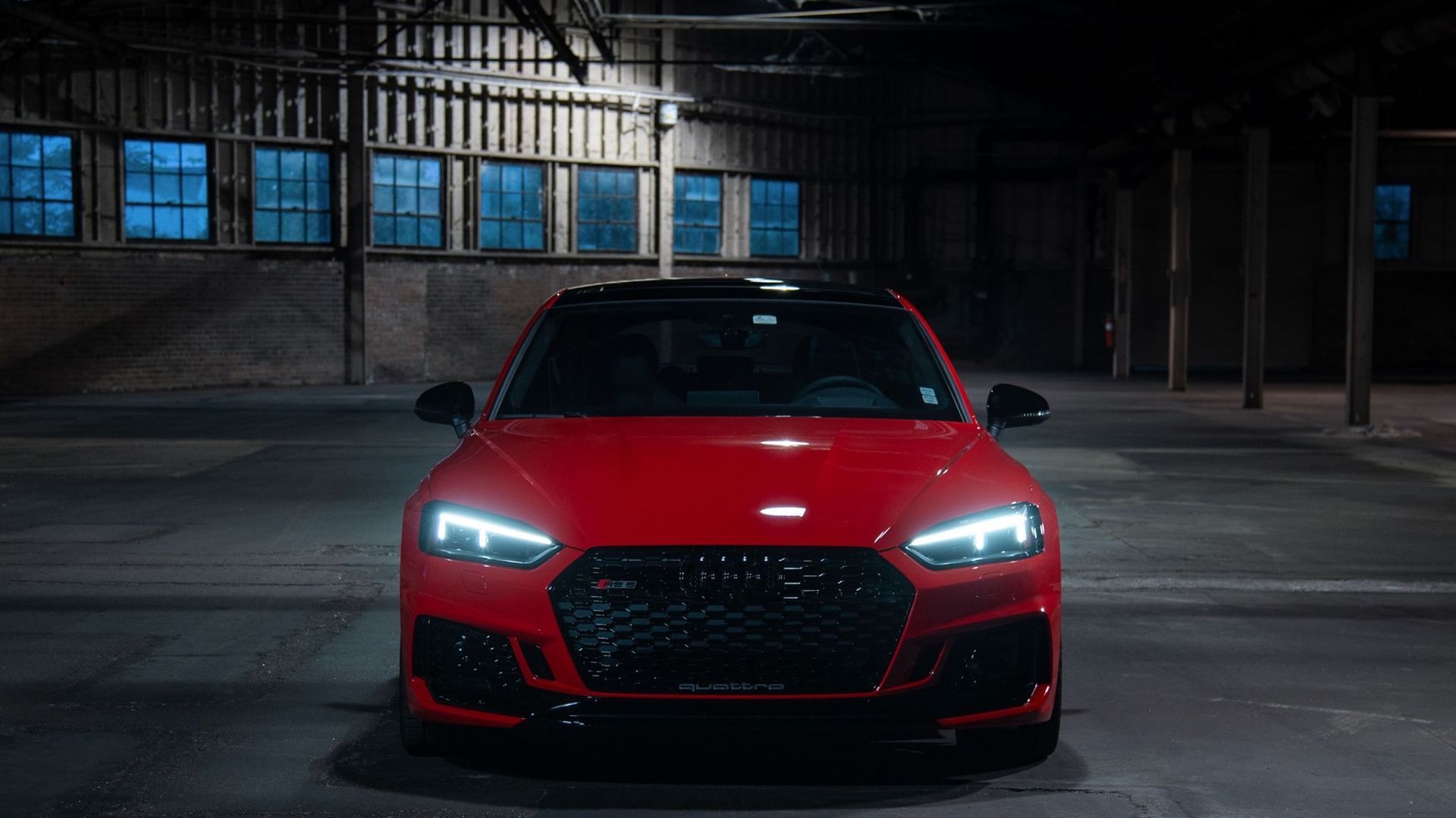 Red Audi sports car parked in a dark, industrial space, headlights on.