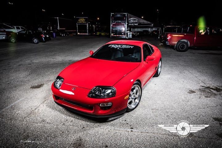 Red Toyota Supra parked on asphalt at night, with other vehicles and trailers in the background.