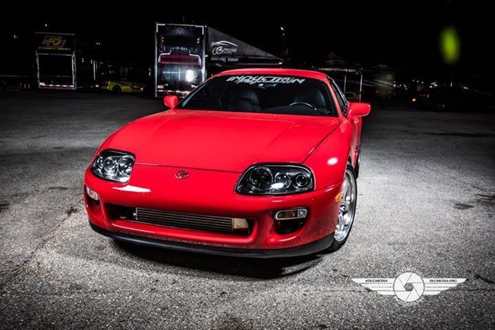 Red Toyota Supra sports car on a race track at night, with headlights on.