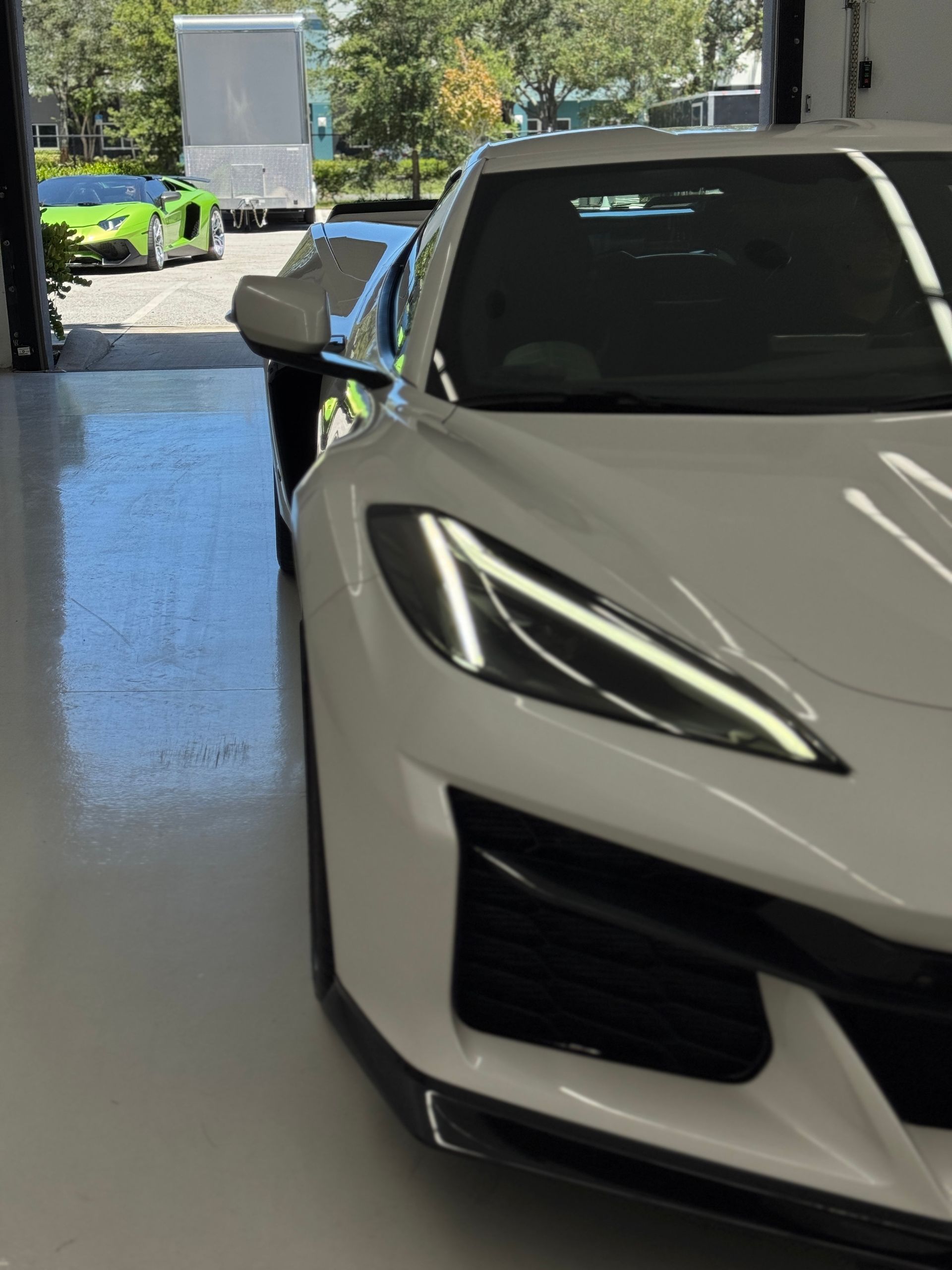 White sports car inside a garage with a view of a green sports car and a truck outside.