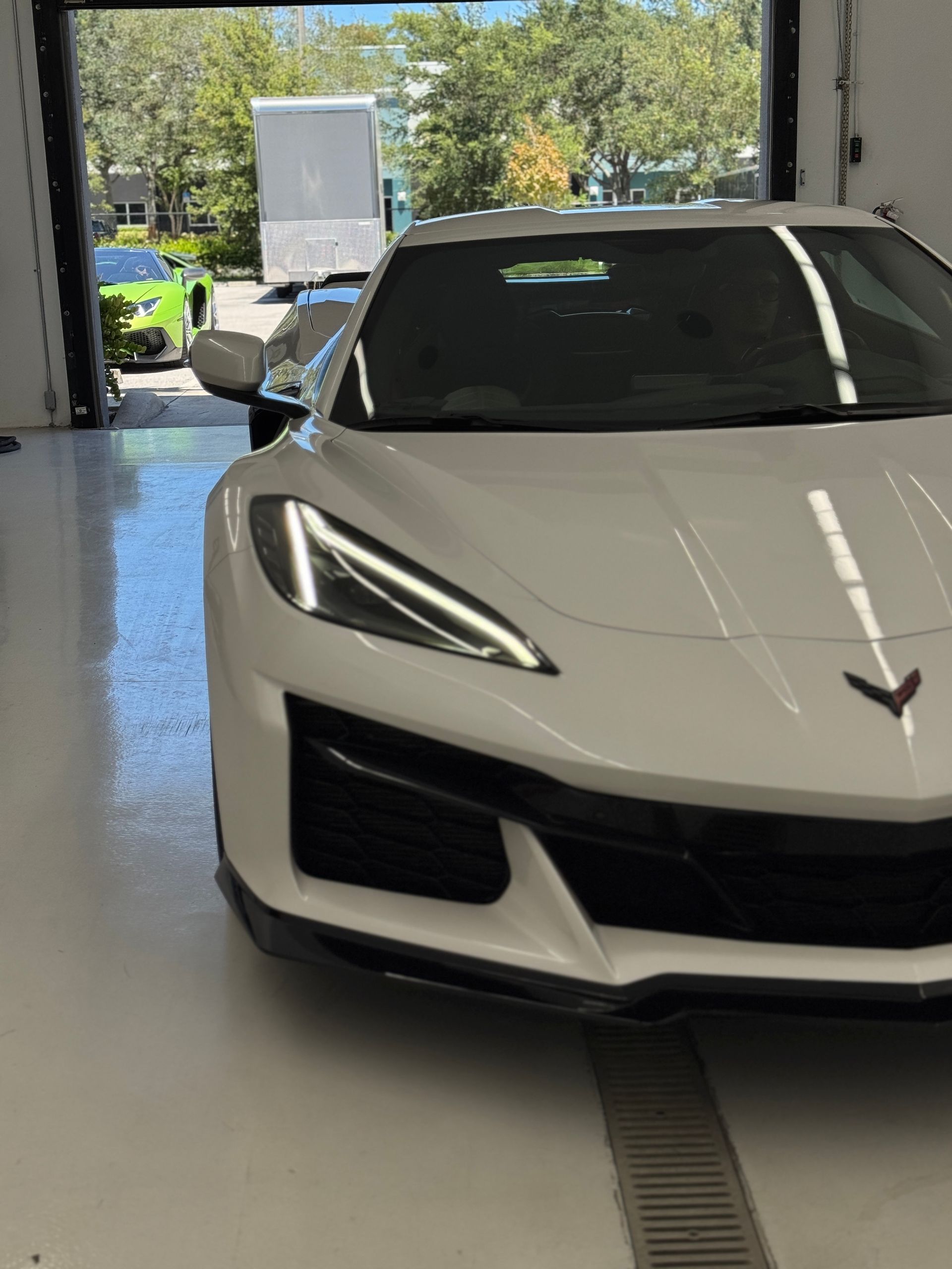 White sports car, partially in a garage, black grille, another green car behind.
