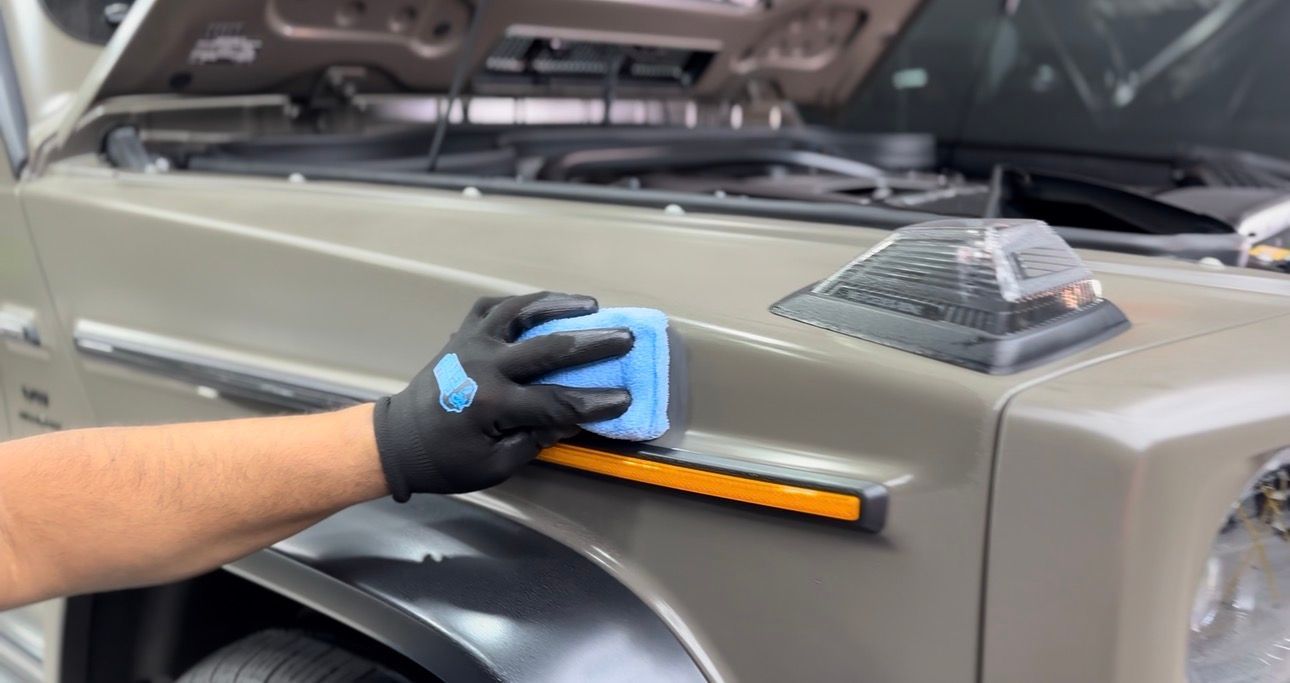 Person applying detailing product to a tan car's side panel with a blue sponge and black gloves.