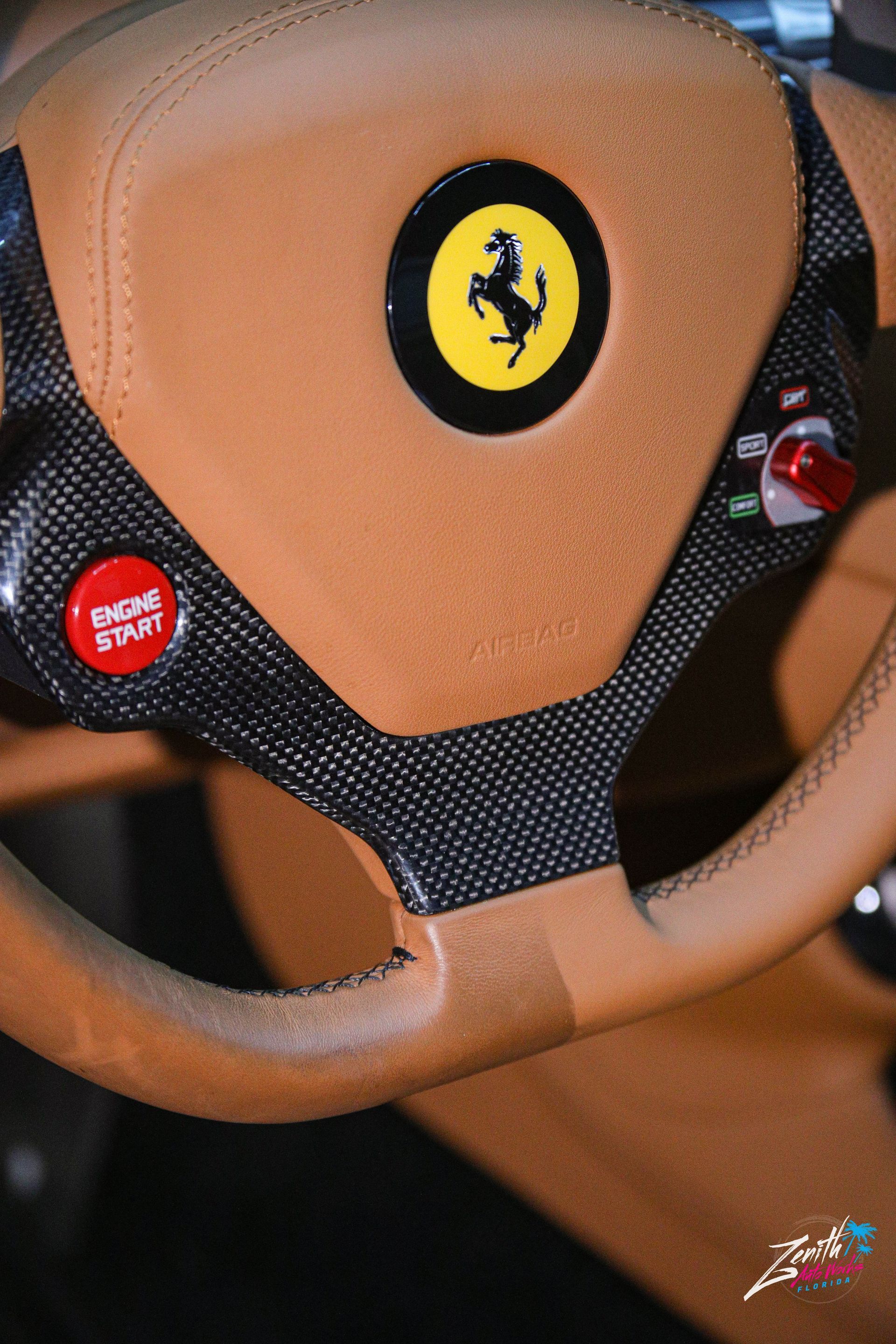 Tan Ferrari steering wheel with carbon fiber accents, featuring the brand's logo and start button.