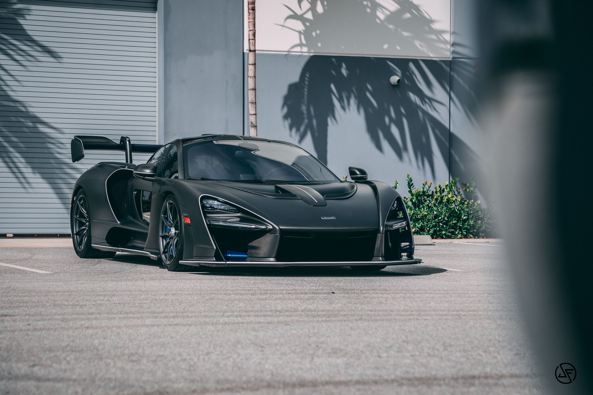 Black McLaren Senna parked outdoors, palm tree shadows on a light-colored building.