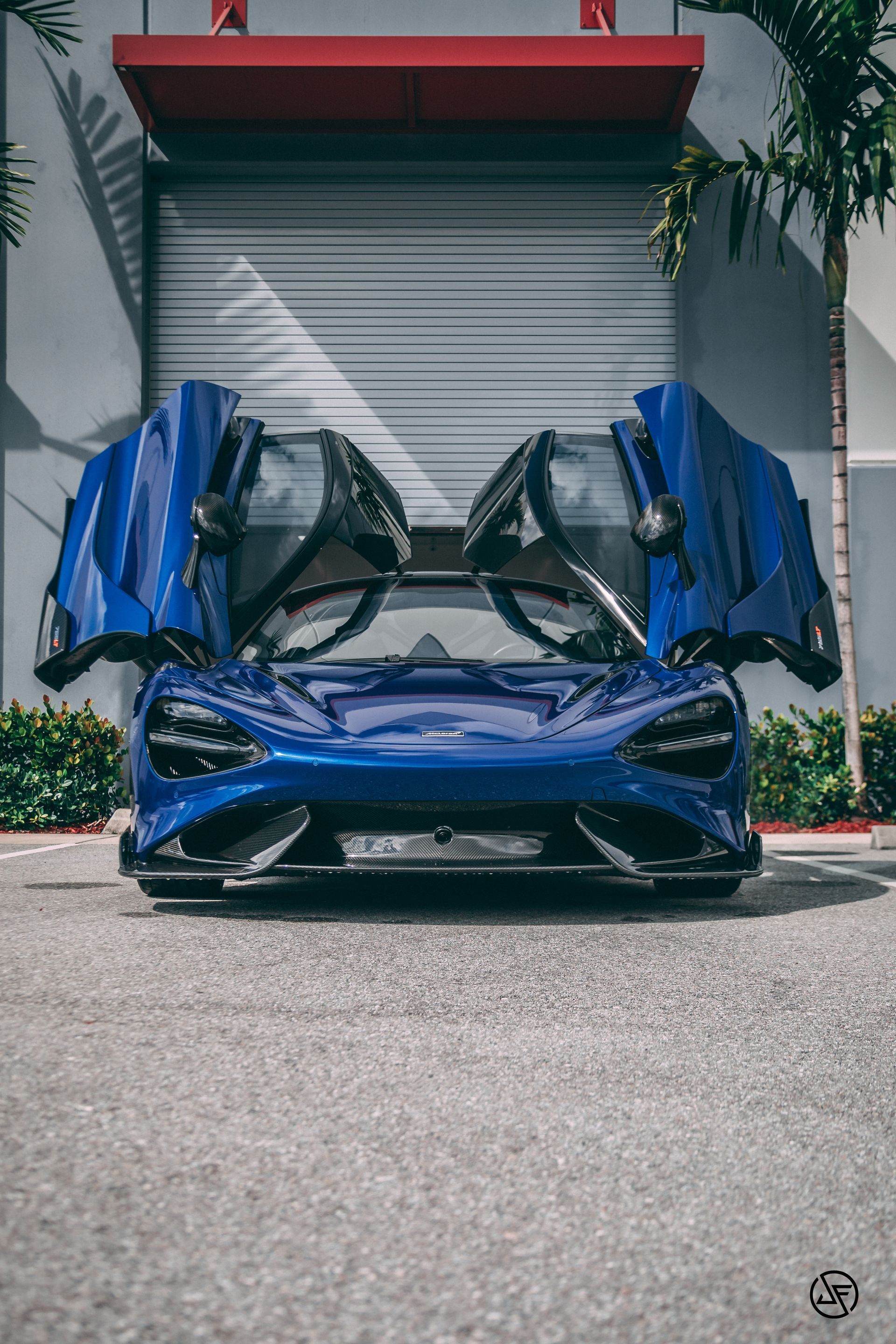 Blue McLaren sports car with doors open in front of a gray garage.