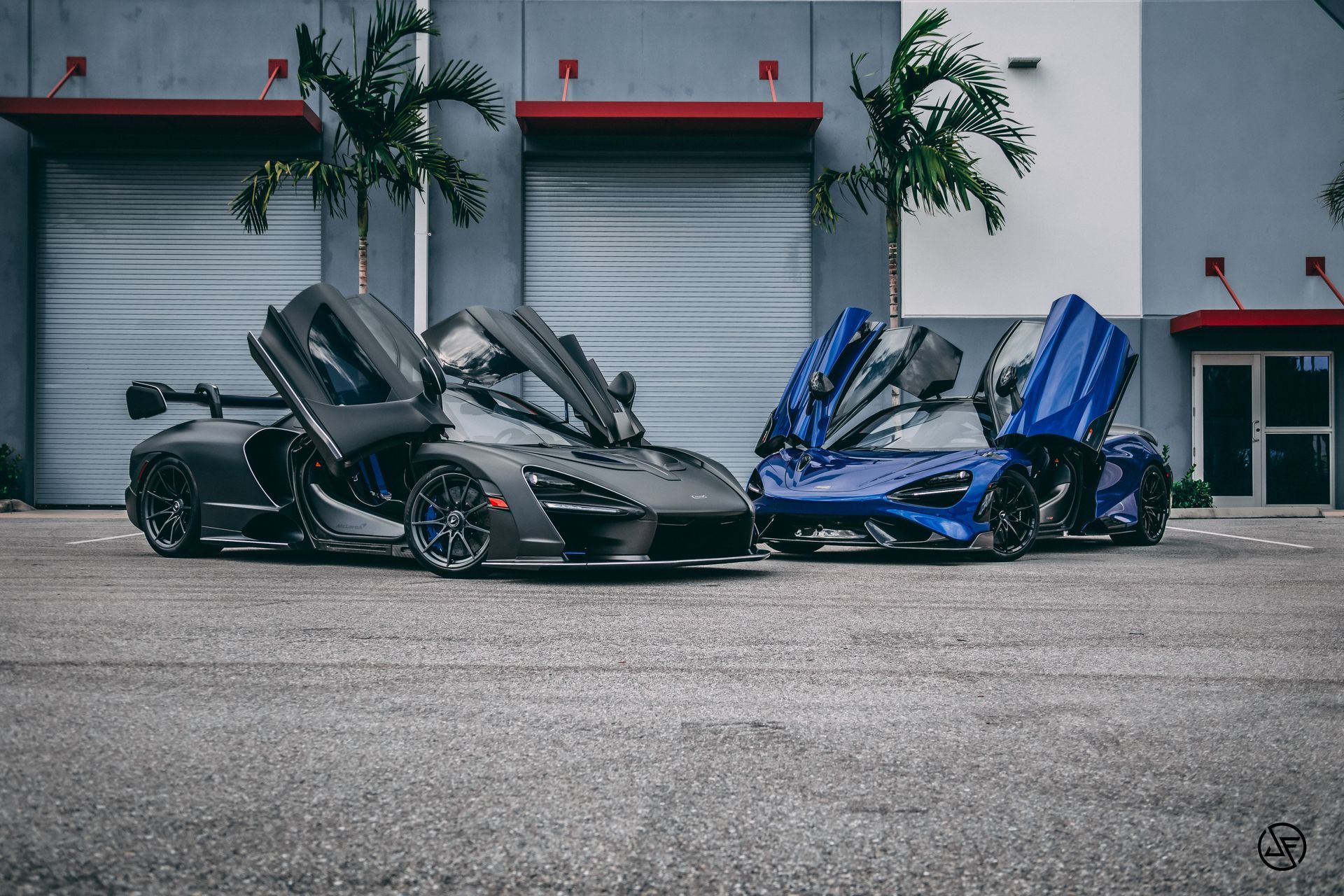Two McLaren supercars with doors up, black and blue, parked in front of industrial building.