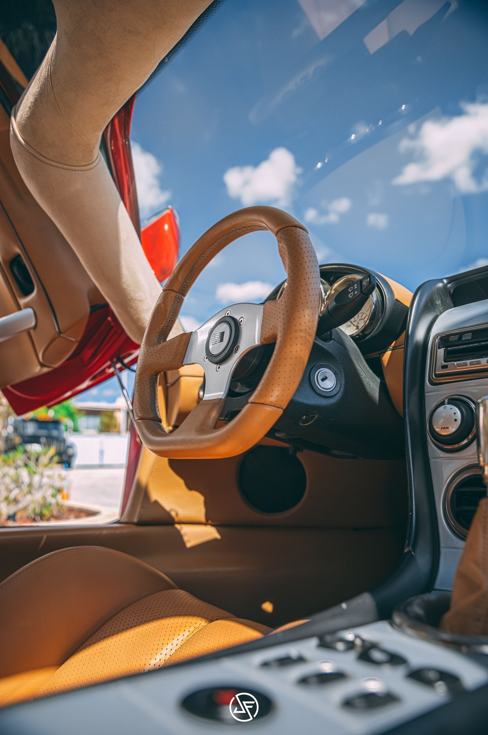 Tan interior of a red sports car. Leather steering wheel, open door, and sunny sky are visible.