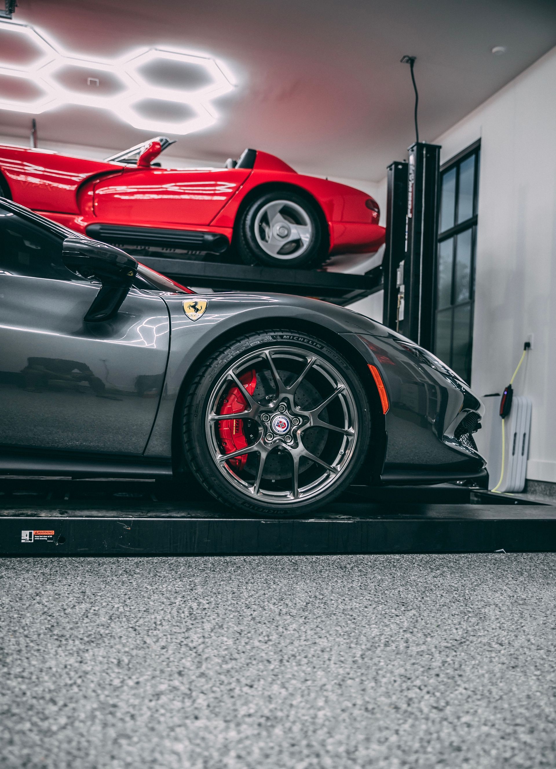 Gray Ferrari and red sports car on a two-post lift in a garage with specialized lighting.