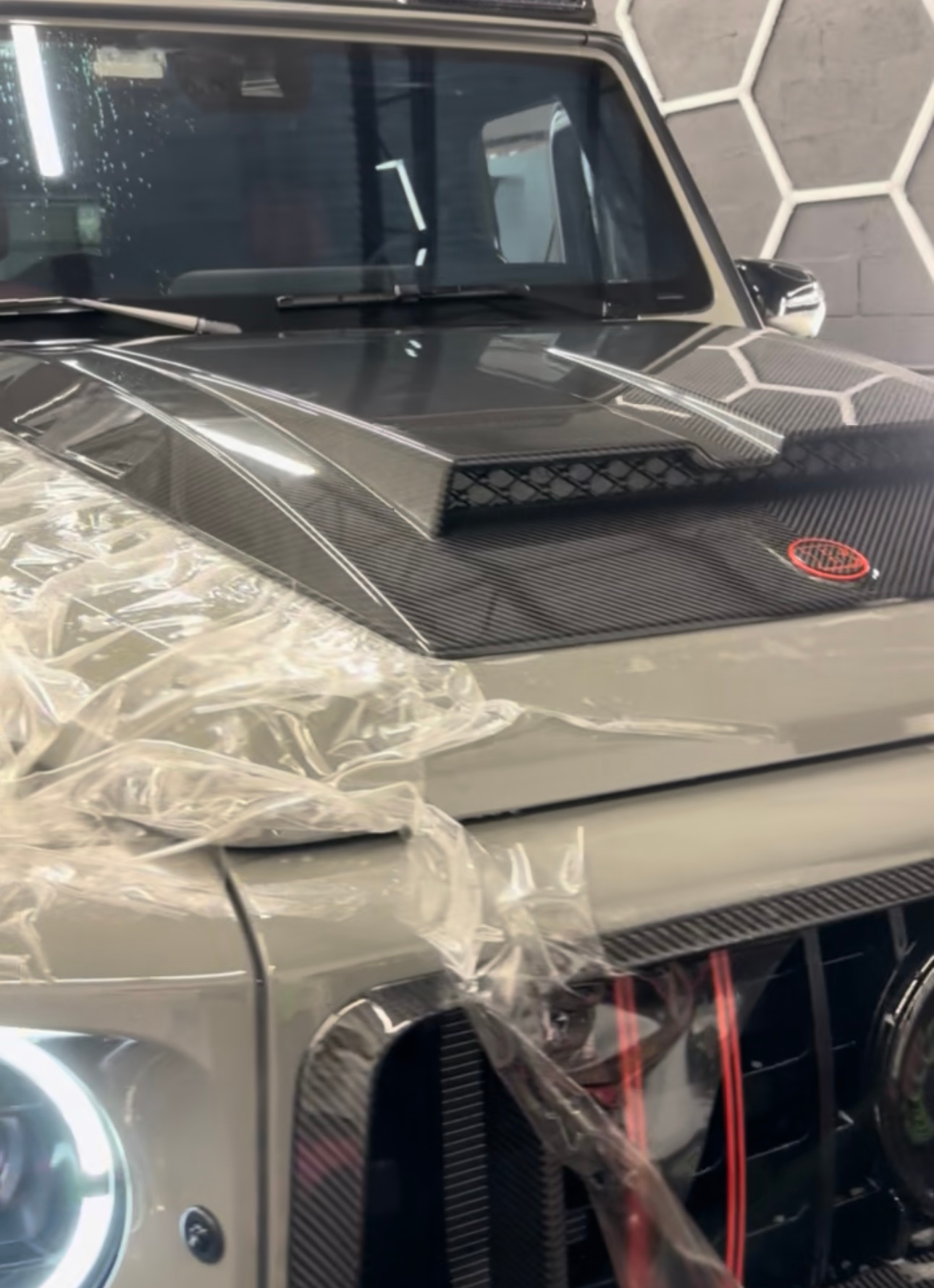 Close-up of a gray Mercedes-Benz G-Class SUV, with a carbon fiber hood and grille, covered in plastic.