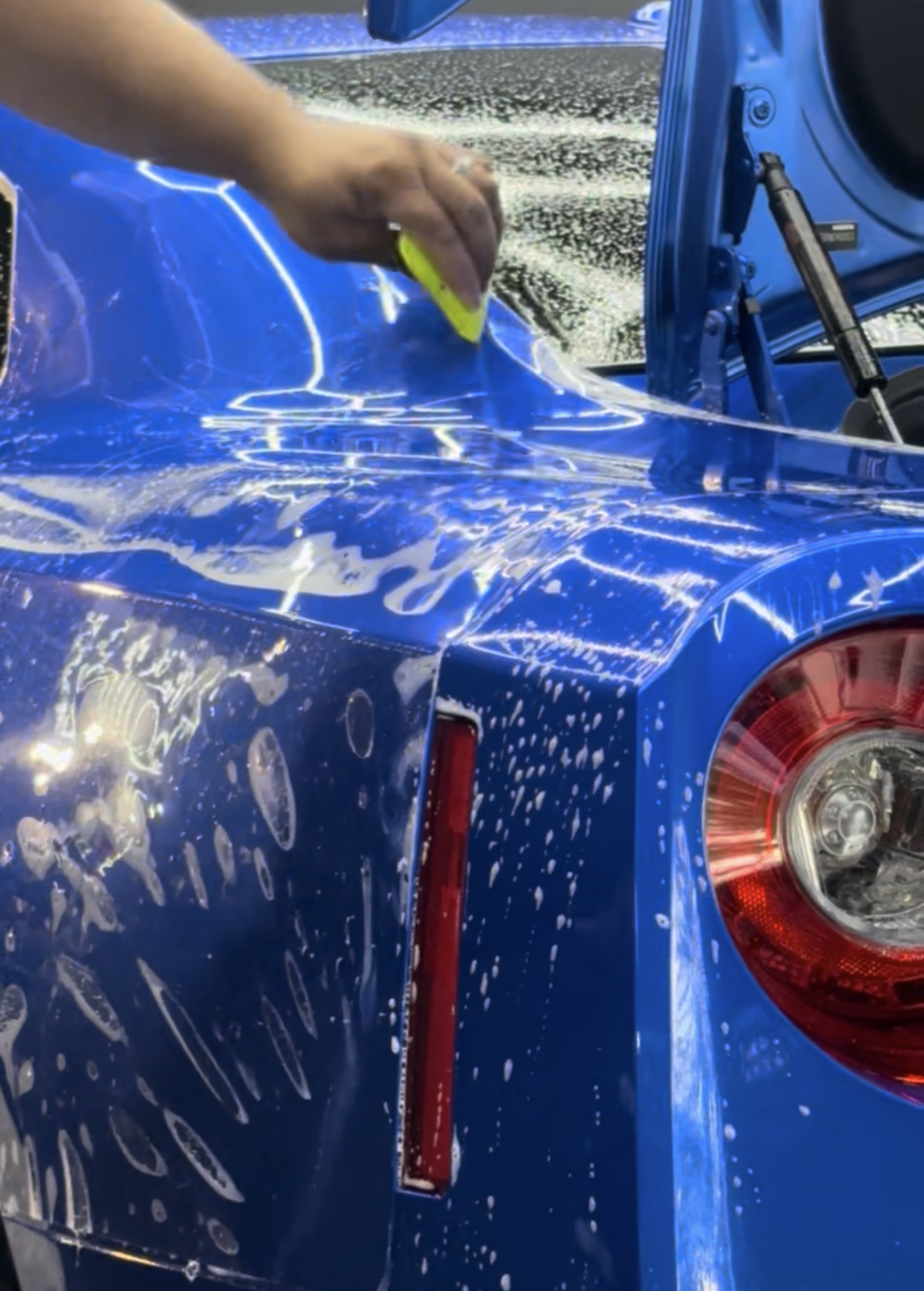 Person uses a squeegee to apply film to a blue car's rear quarter panel, covered in soapy water.