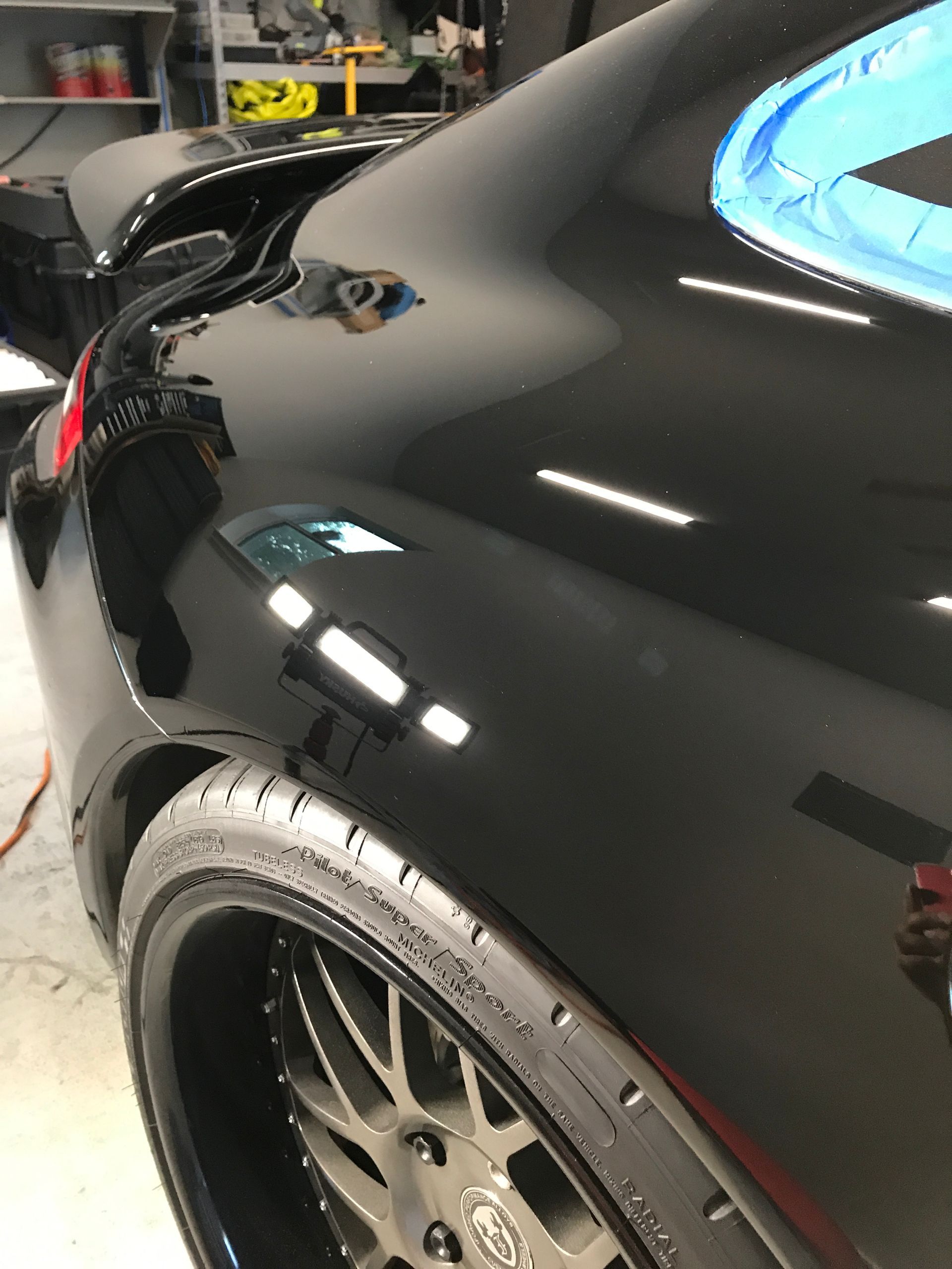 Black car's glossy finish; rear quarter panel, tire, and wheel are in focus. Blue tape lines edges.