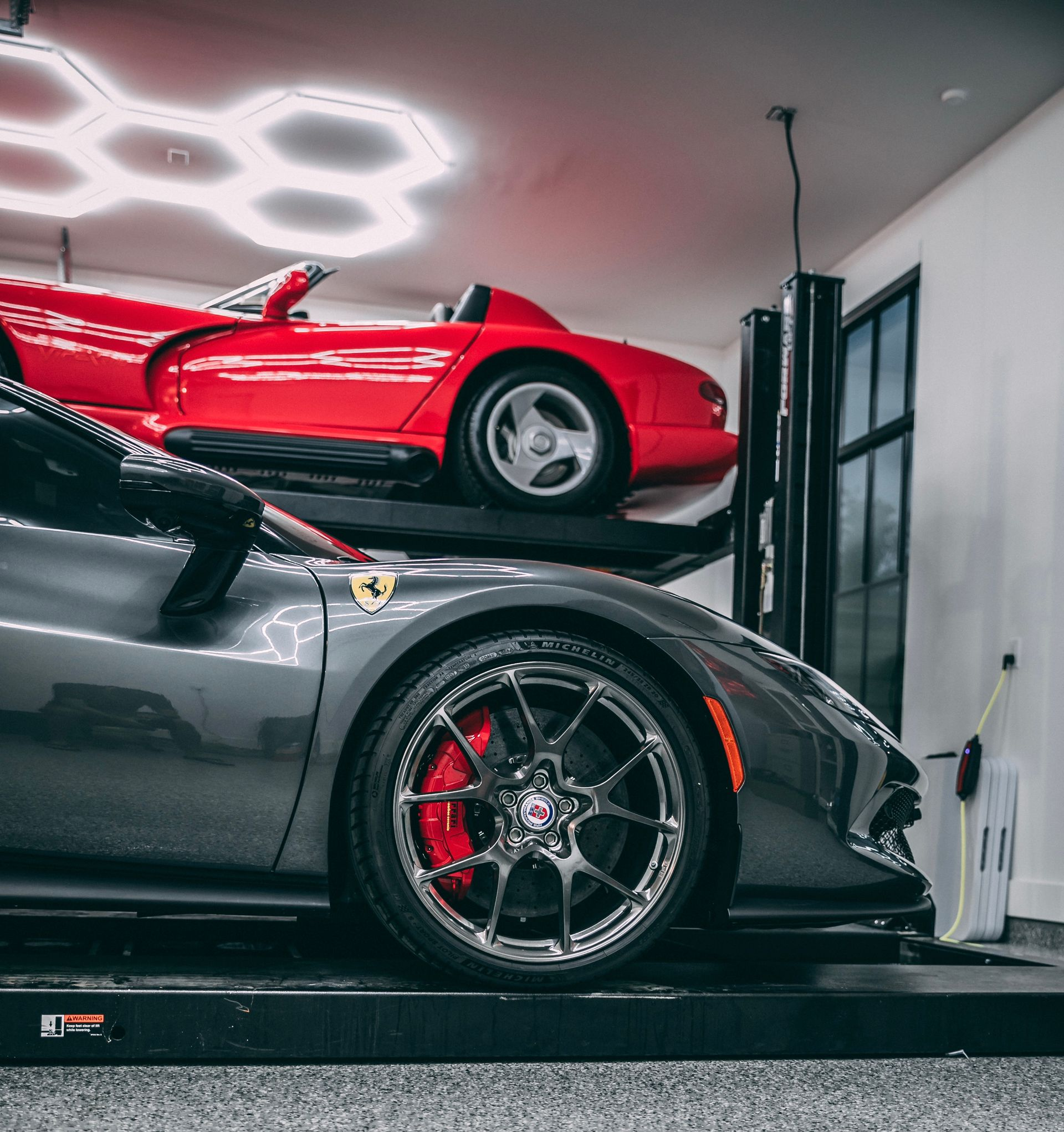 Gray Ferrari and red Dodge Viper on a car lift in a garage with honeycomb lights.