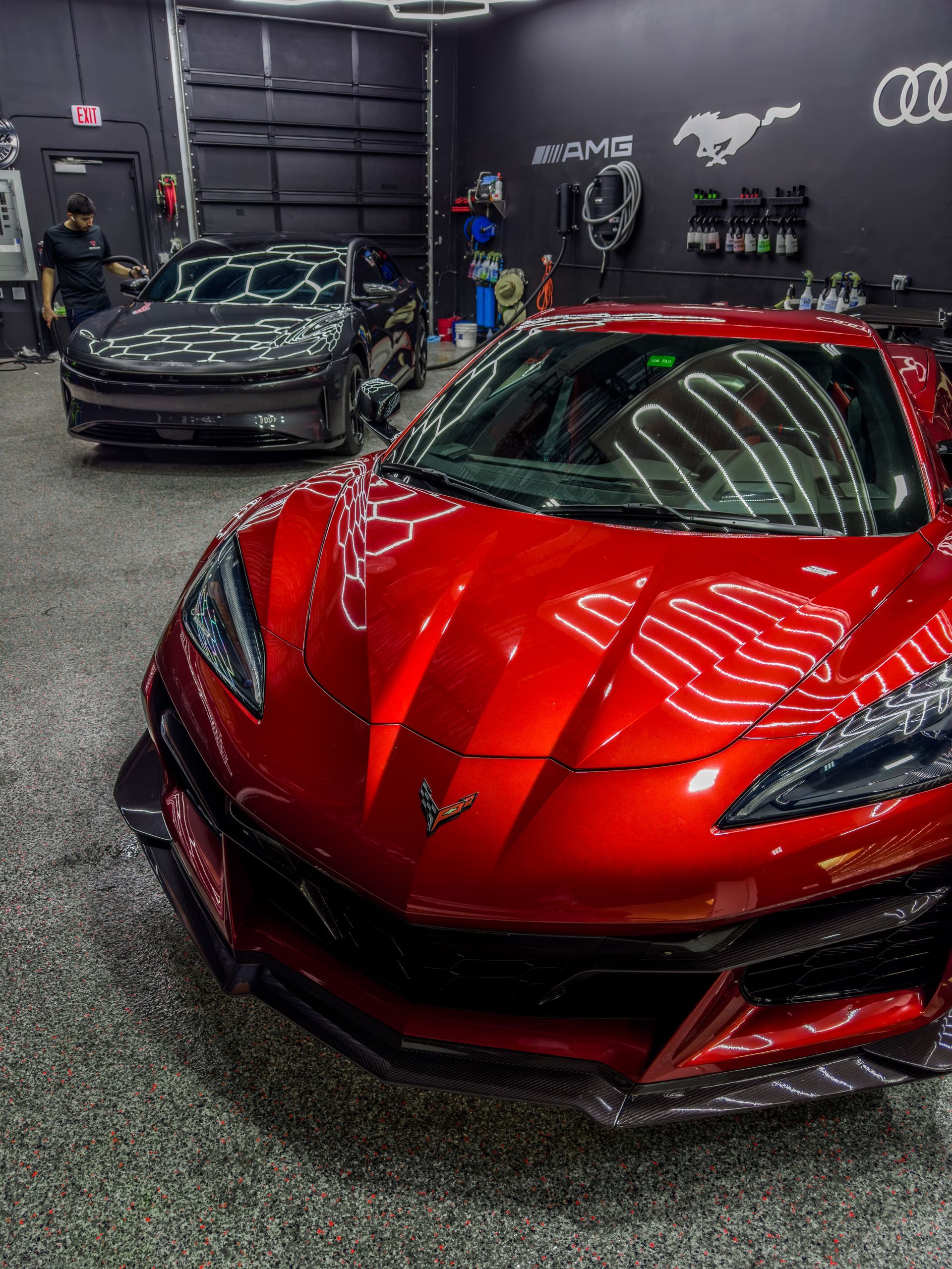 Red sports car in a garage, another car in background. Shiny red paint, grey floor, shop setting.