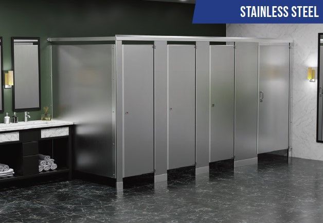 Fila de cubículos de baño de acero inoxidable en un baño público con piso de baldosas oscuras y lavabos a la izquierda.