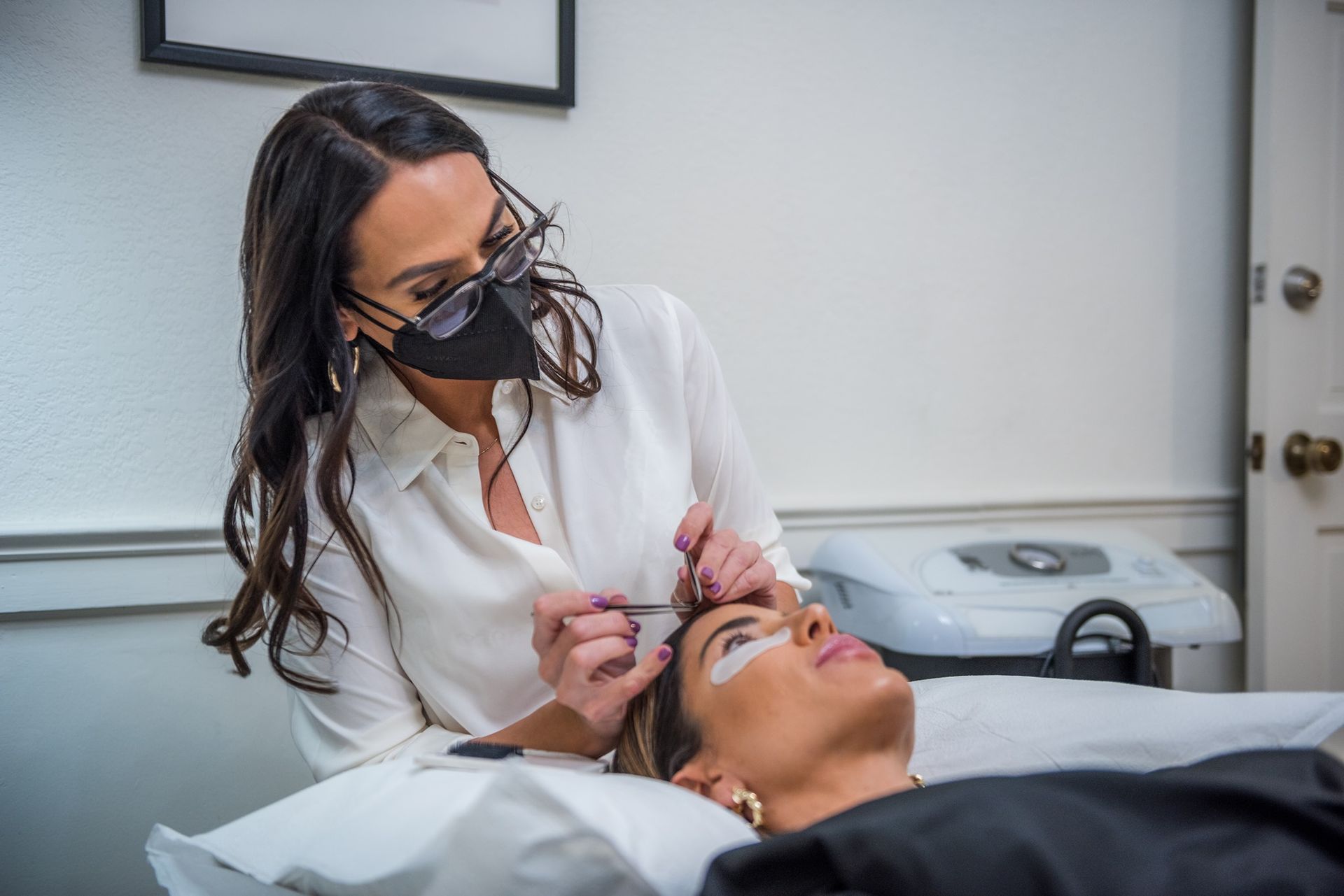 A woman wearing a mask is applying eyelash extensions to a woman.