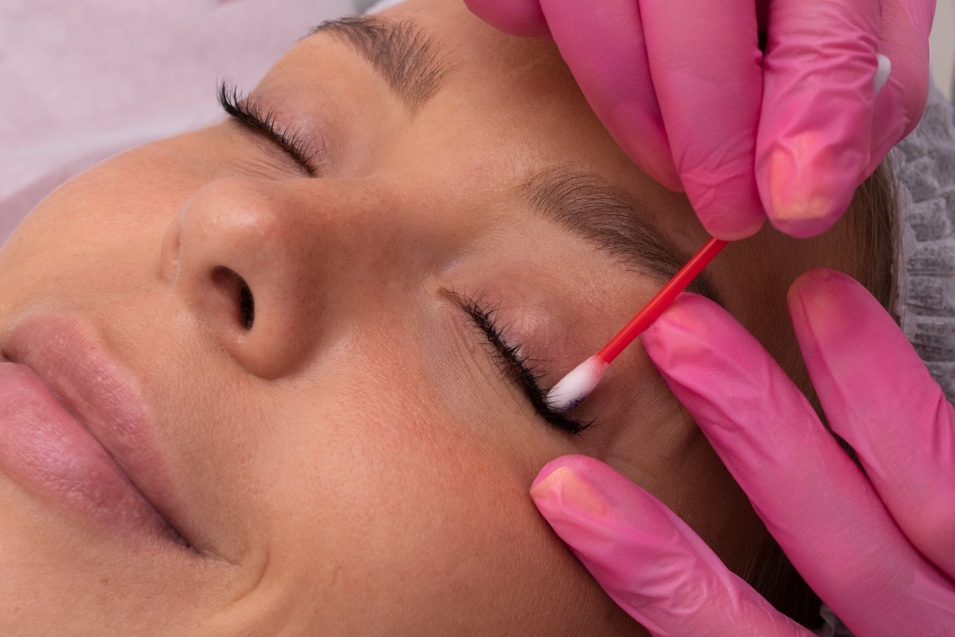 A woman is getting her eyebrows done by a woman wearing pink gloves.