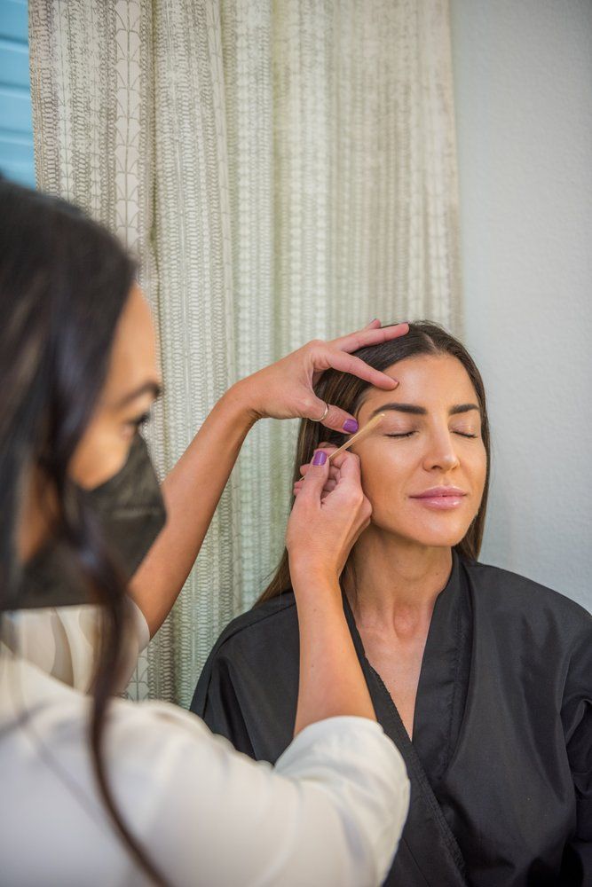 A woman is getting her eyebrows done by a makeup artist while wearing a mask.