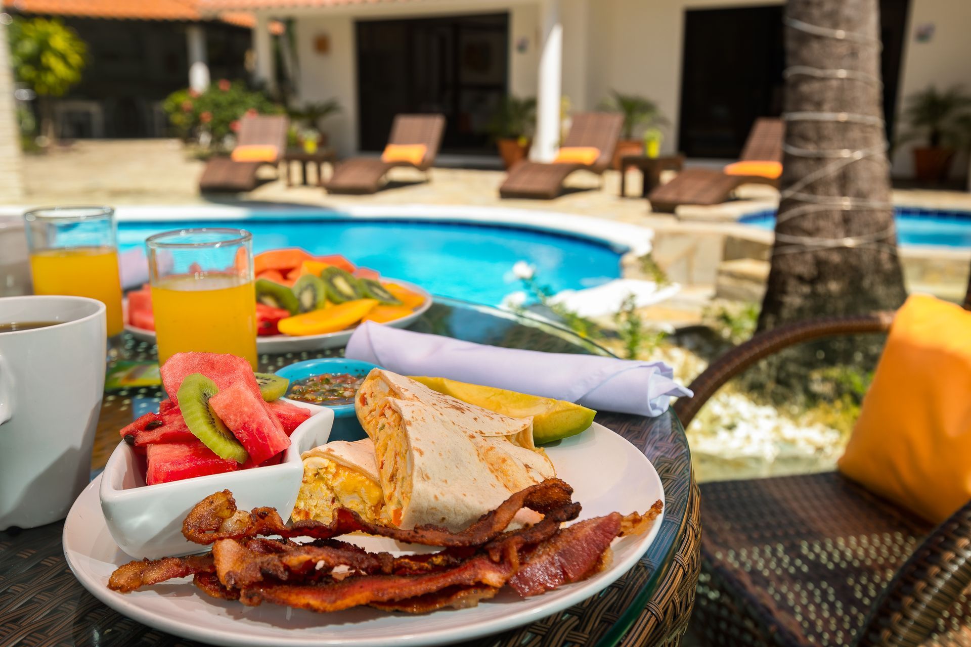 Breakfast plate with breakfast burrito, bacon, fruit, and juice, by a pool with lounge chairs and the trunk of a palm tree.
