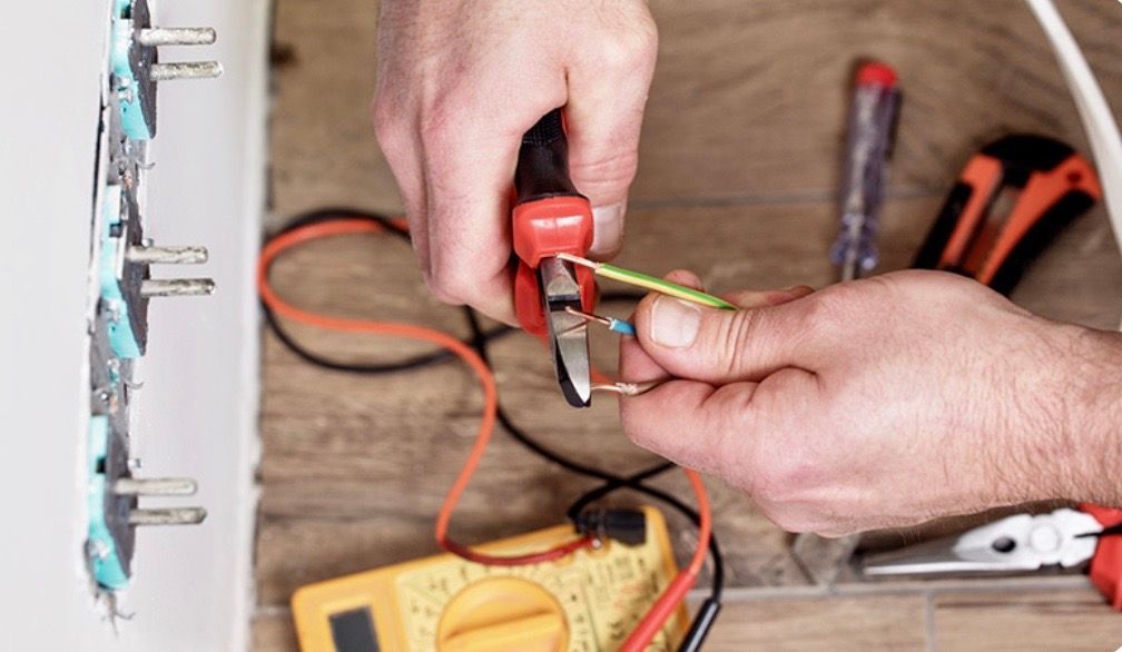 Electrician cutting a wire with pliers near a wall outlet, with a multimeter and tools nearby.