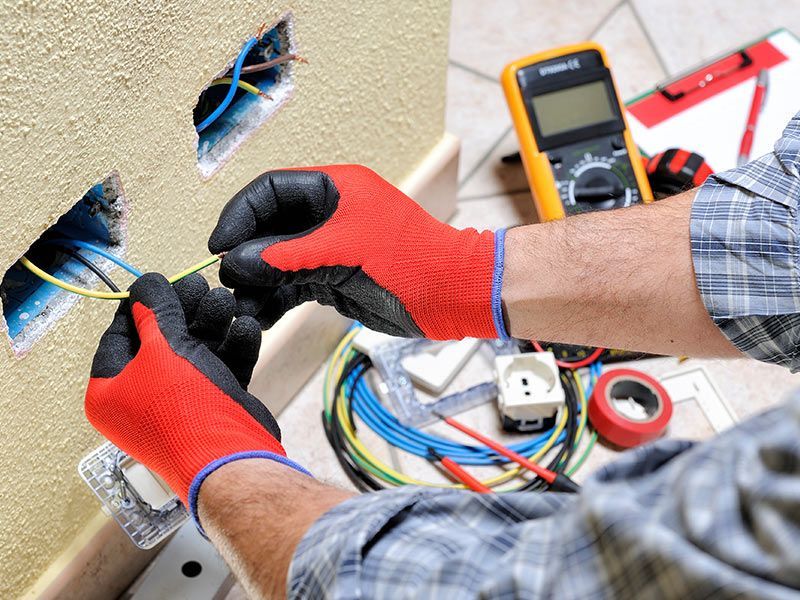 Electrician in red gloves wiring electrical outlets on a wall. Yellow multimeter and other tools present.