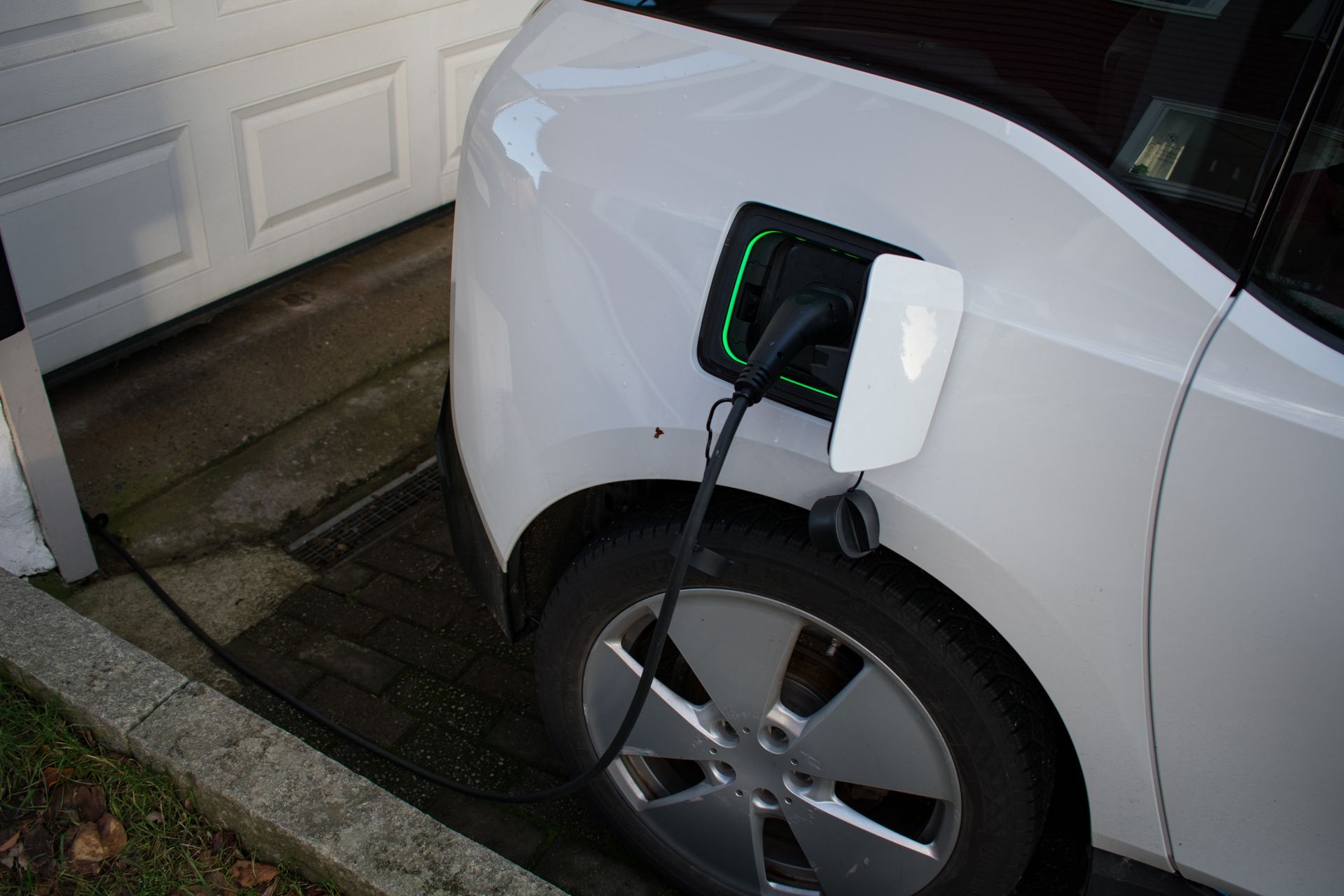 A white electric vehicle parked near a garage with a charging cable plugged into its side port.