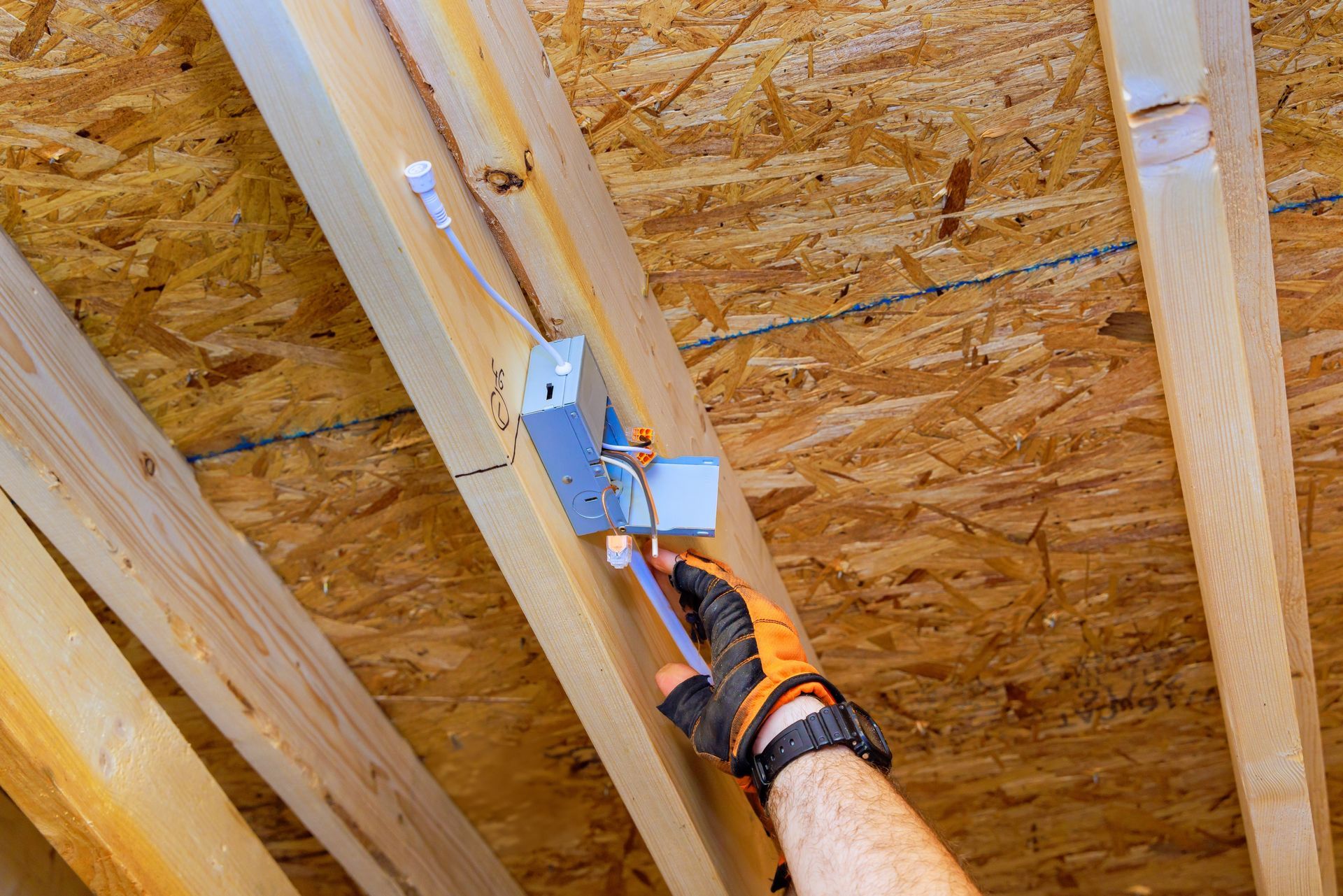 Hand with orange glove wiring electrical box in attic with wooden beams.