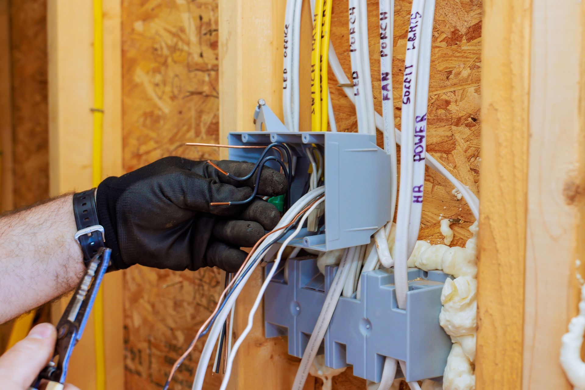 A gloved hand uses pliers to strip insulation from wires inside a gray electrical junction box mounted on wood framing.