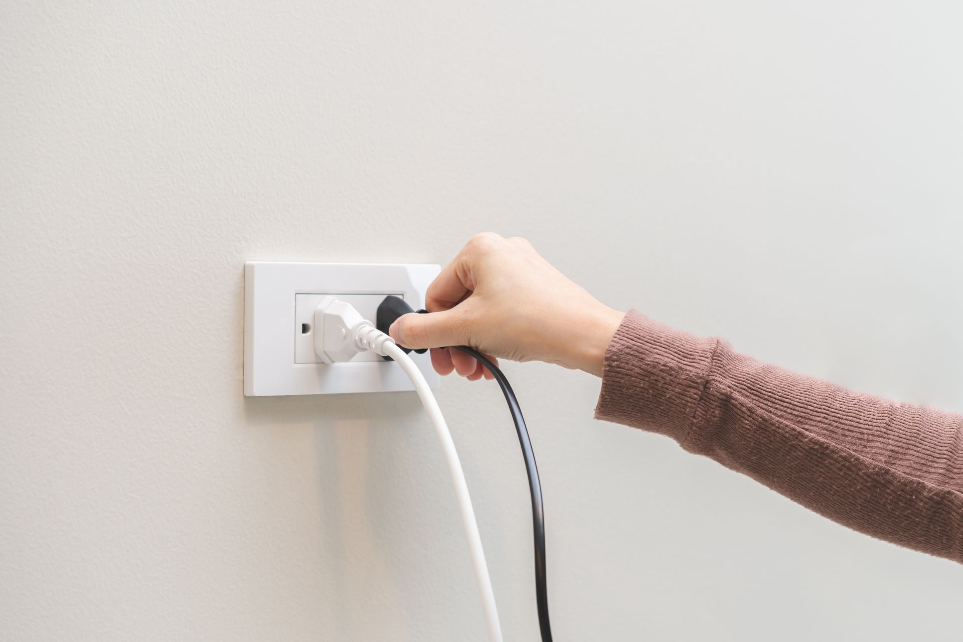 A hand in a brown sleeve unplugging a black power cord from a white wall outlet that already has a white cord plugged in.