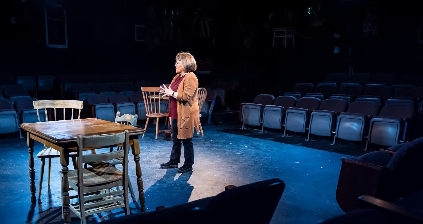 A woman is standing on a stage in front of a table and chairs.